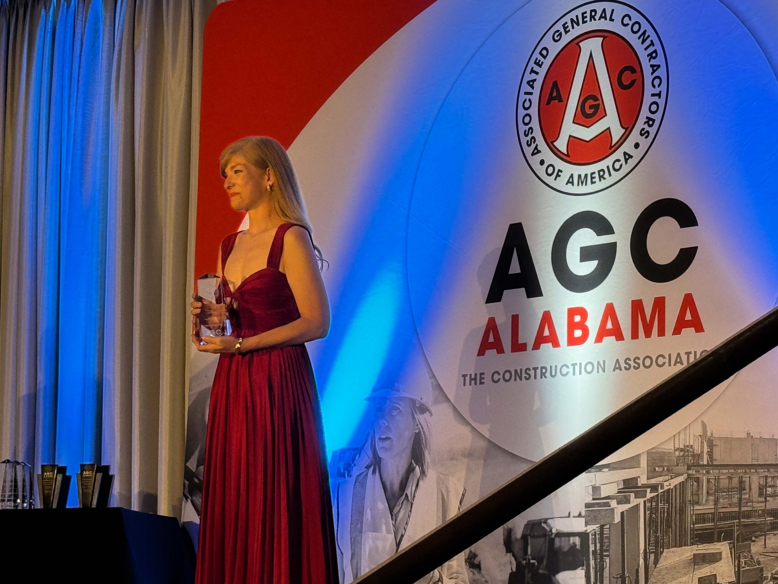 A woman in a long red dress holding an award standing on a stage next to a large banner for the Associated General Contractors of America Alabama chapter, with a construction site image in the background.