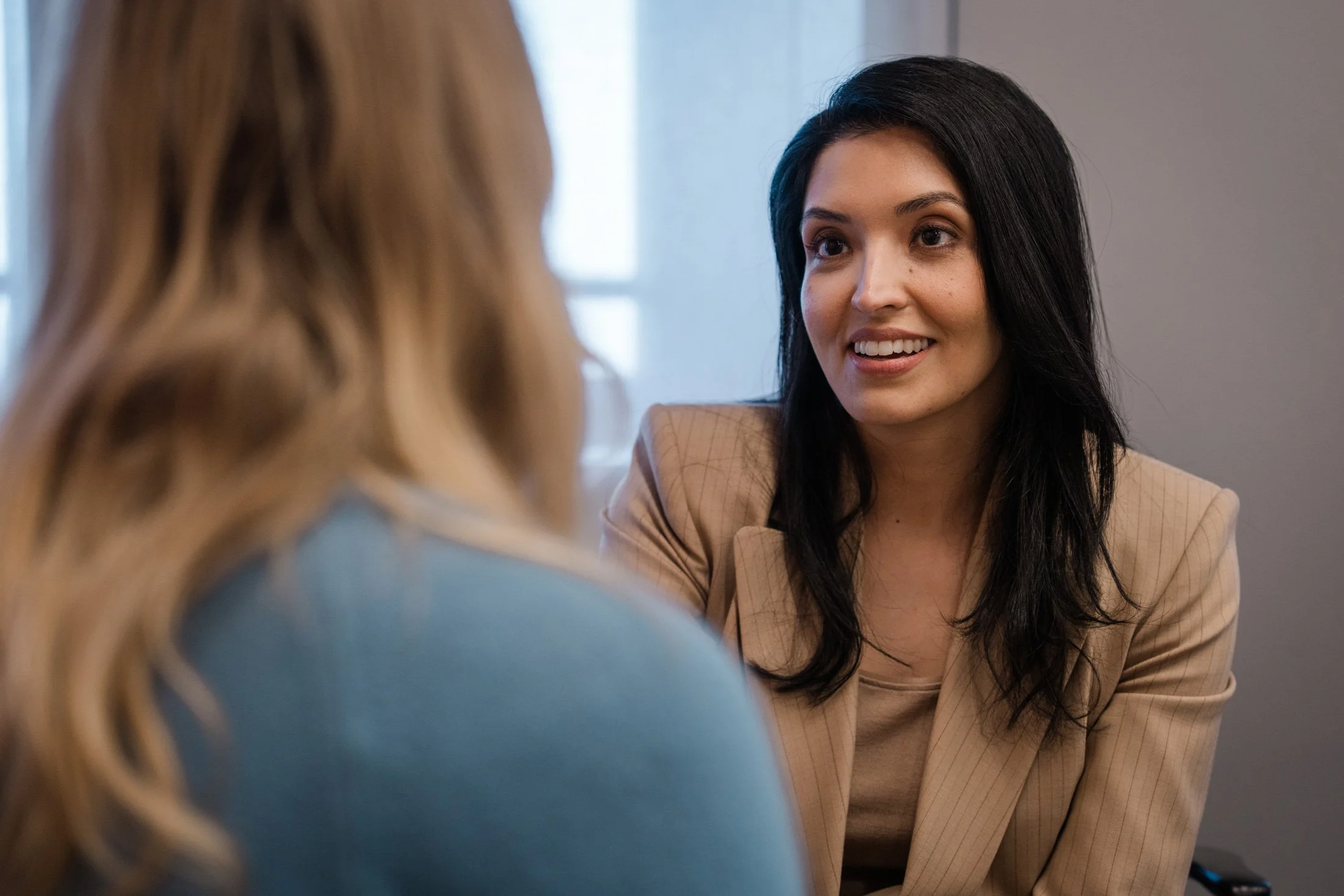 A woman with long black hair and a beige blazer smiling during a conversation, with part of another woman with blonde hair visible in the foreground.