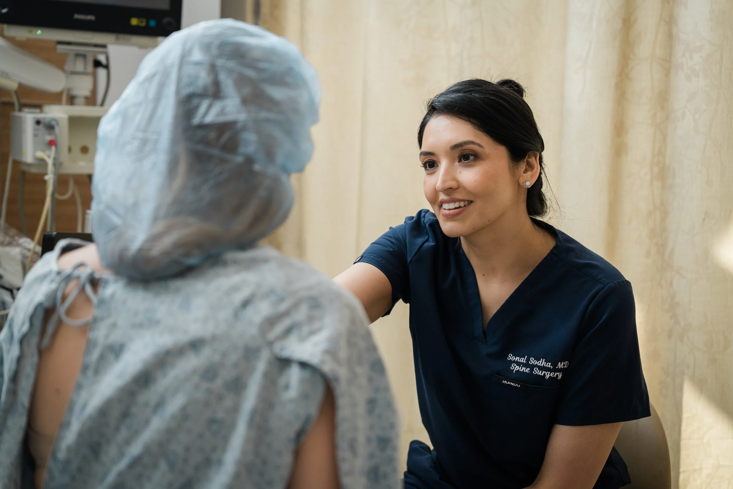 A healthcare professional in dark scrubs smiling and talking to a patient with a bandaged arm in a medical setting.