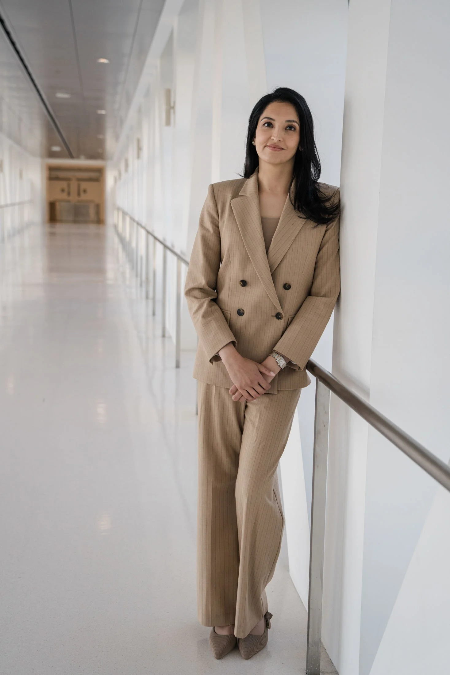 A woman with dark hair is leaning against a white wall in a modern, bright corridor. She is wearing a beige pinstripe suit and beige shoes.
