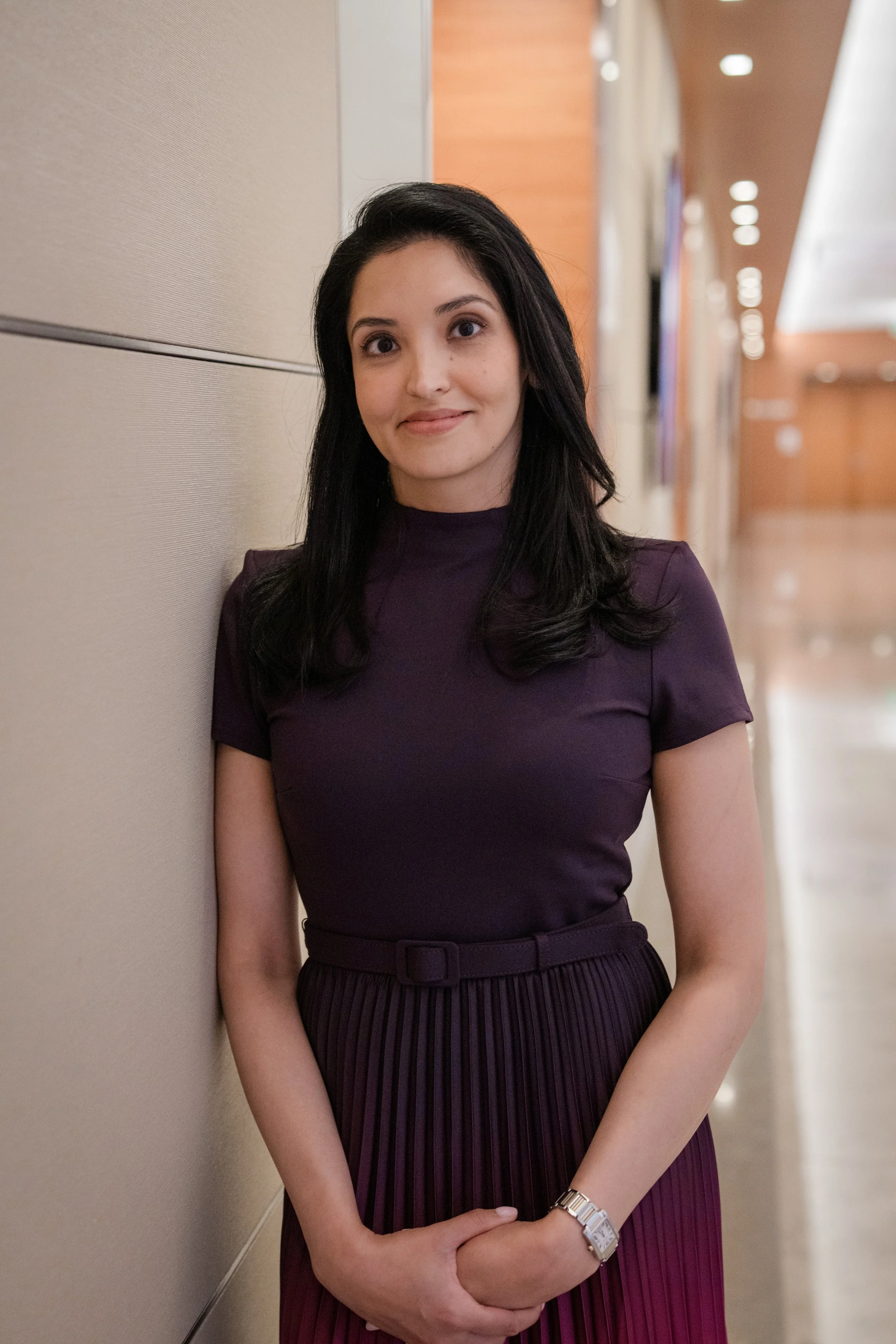 A woman with dark hair in a purple top and pleated skirt standing in a hallway, leaning against a beige wall, smiling softly.