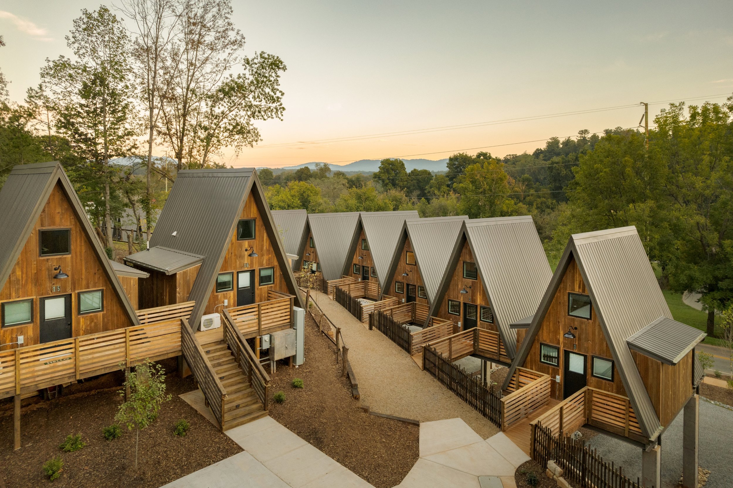 A row of A-frame cabins with wooden exteriors and metal roofs, each with small decks and stairs, set in a green, wooded area with a road nearby at sunset.