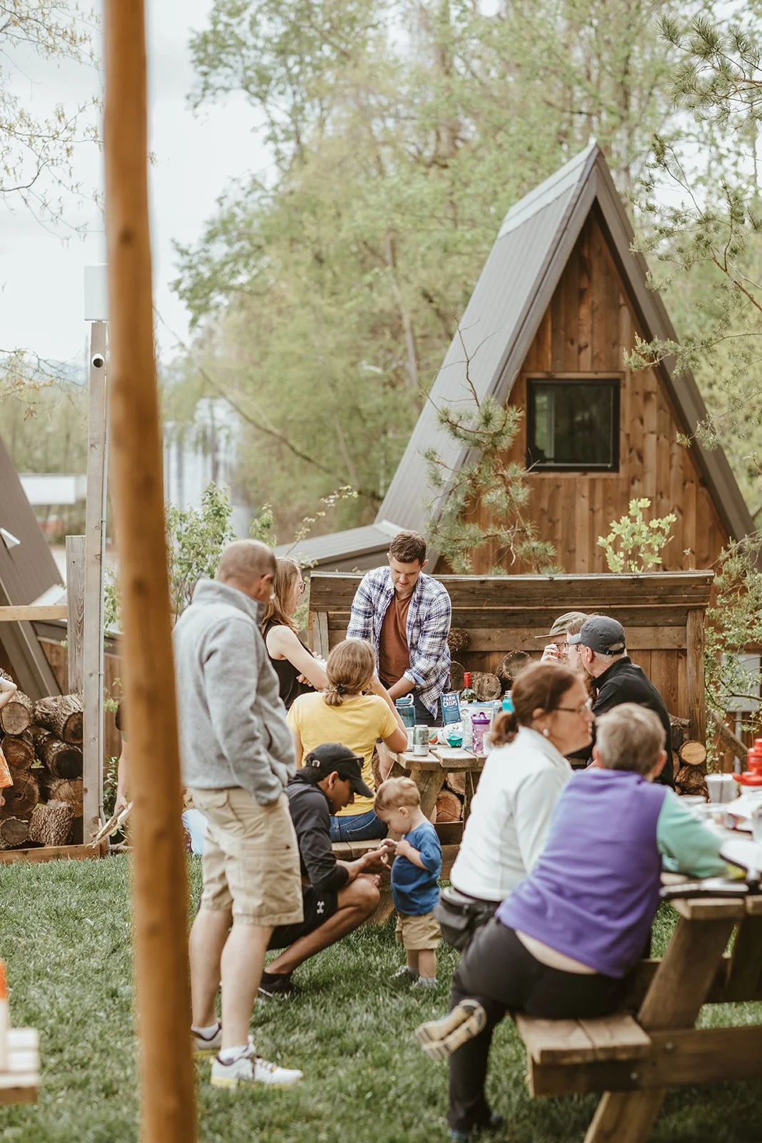 Group of people having a gathering outdoors near a small wooden house with a steep roof, surrounded by trees.