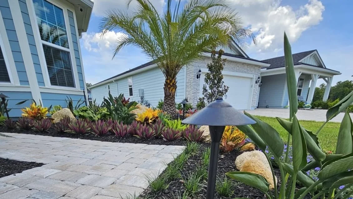 A landscaped front yard with a light-colored brick pathway, colorful plants, a small palm tree, and a modern house with light blue siding and large windows.