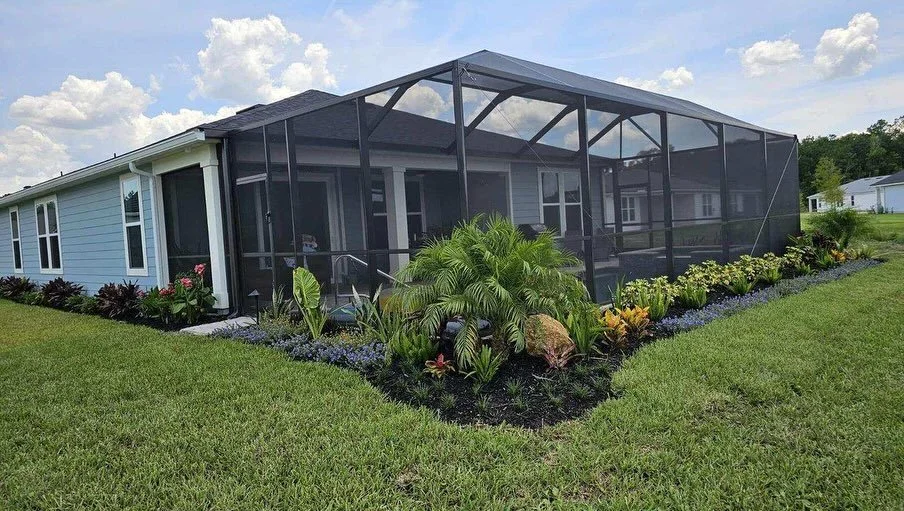 A house with a screened-in porch or patio area, surrounded by a well-maintained lawn and garden with various plants and flowers under a partly cloudy sky. St. Augustine Landscape Design. Builder Grade