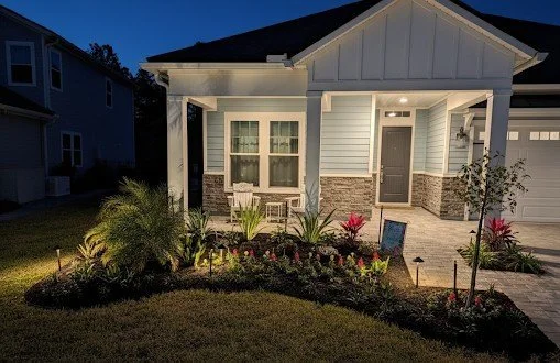 Front view of a house with a well-lit porch, blue exterior walls, white trim, and a small garden with plants and flowers in the evening.