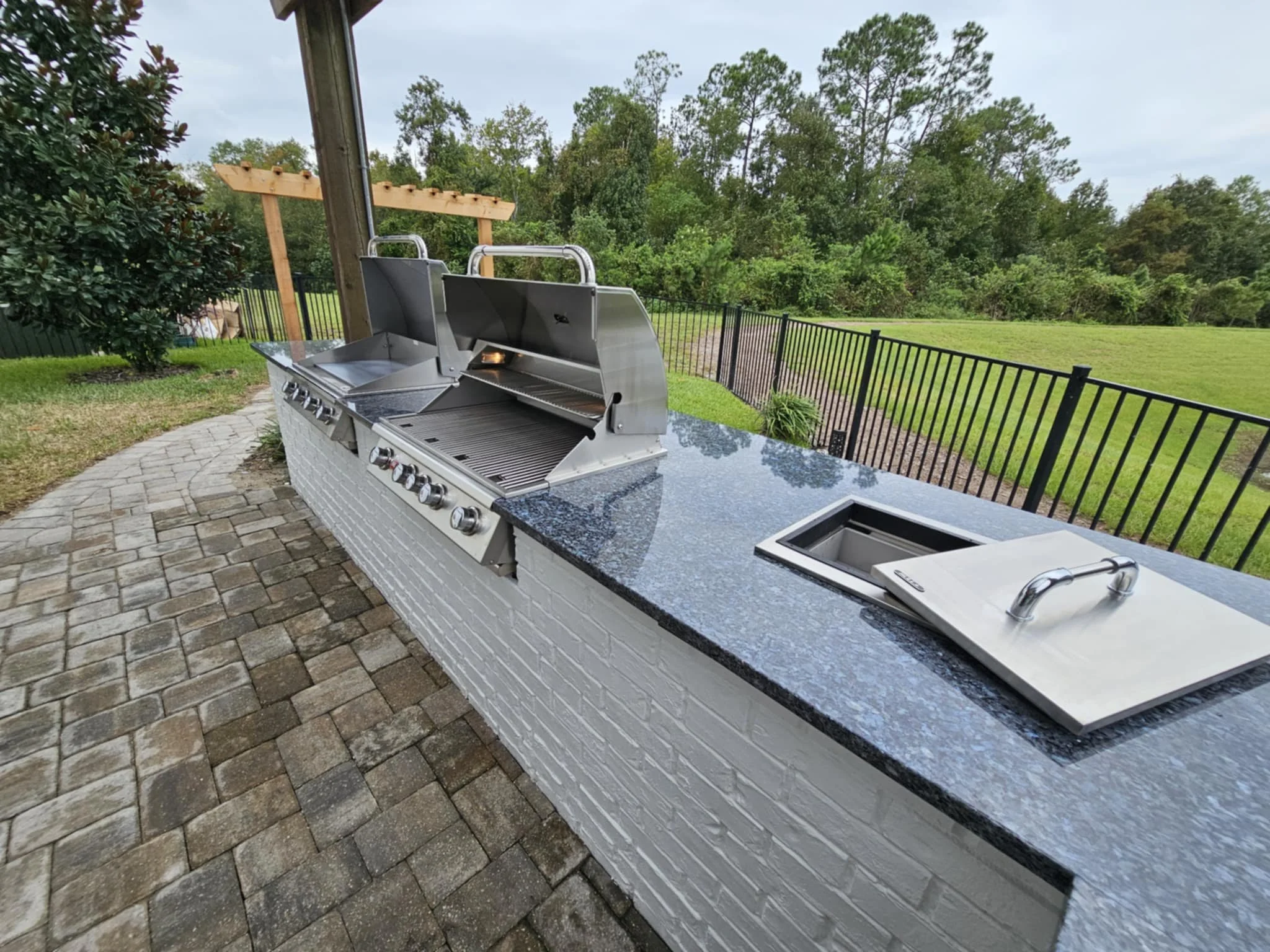 An outdoor kitchen area featuring a built-in stainless steel gas grill with multiple control knobs, a black granite countertop, and a built-in smoker box with a cover. Jacksonville Outdoor Kitchen Contractor.