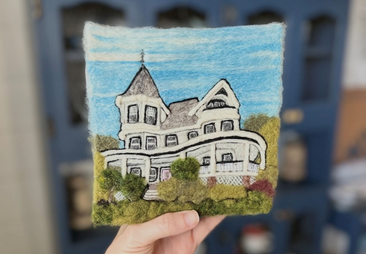 A felted art piece depicting a Victorian-style house with a porch and a tower, featuring a blue sky background.
