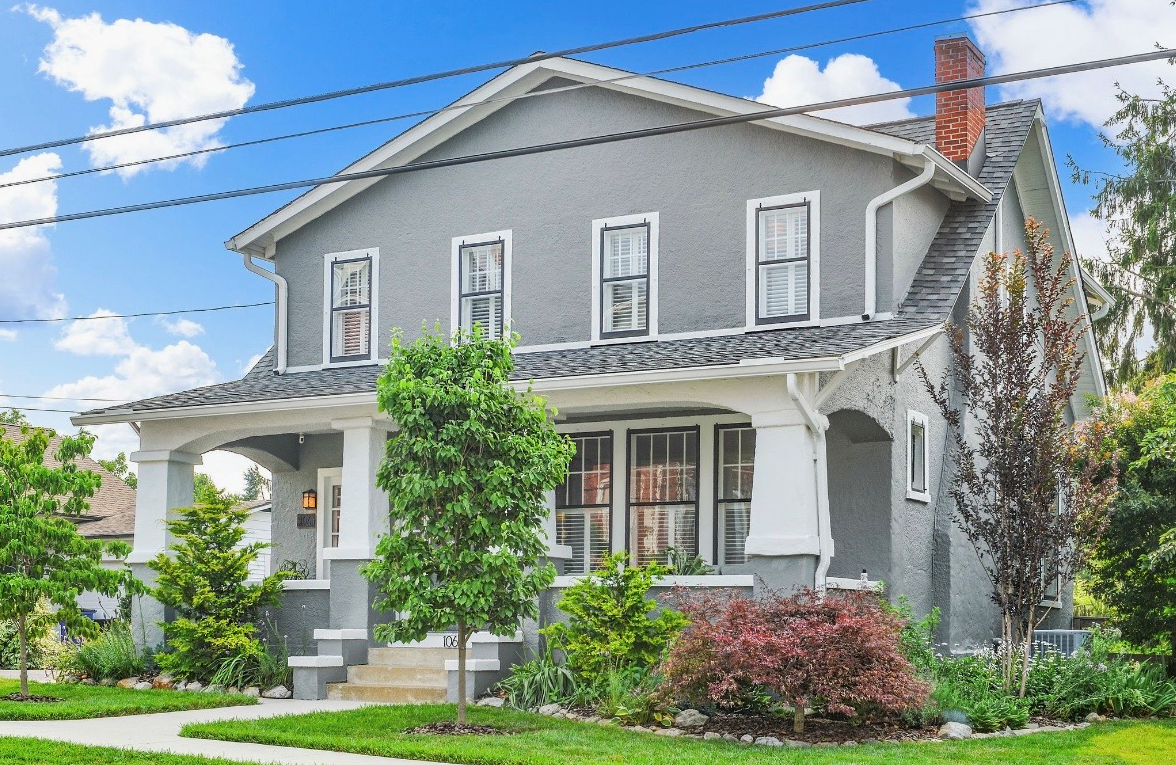 Gray two-story house with front porch, surrounded by green trees and plants, under a bright blue sky with white clouds.