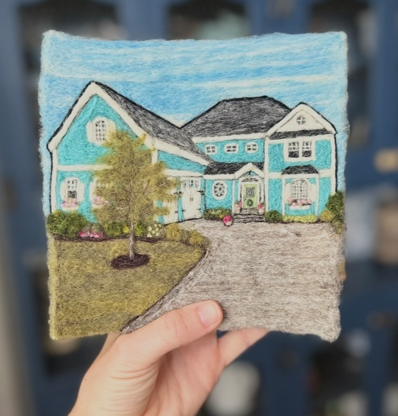 Colorful felt artwork of a blue house with a driveway and landscaped yard, with mountains in the background.