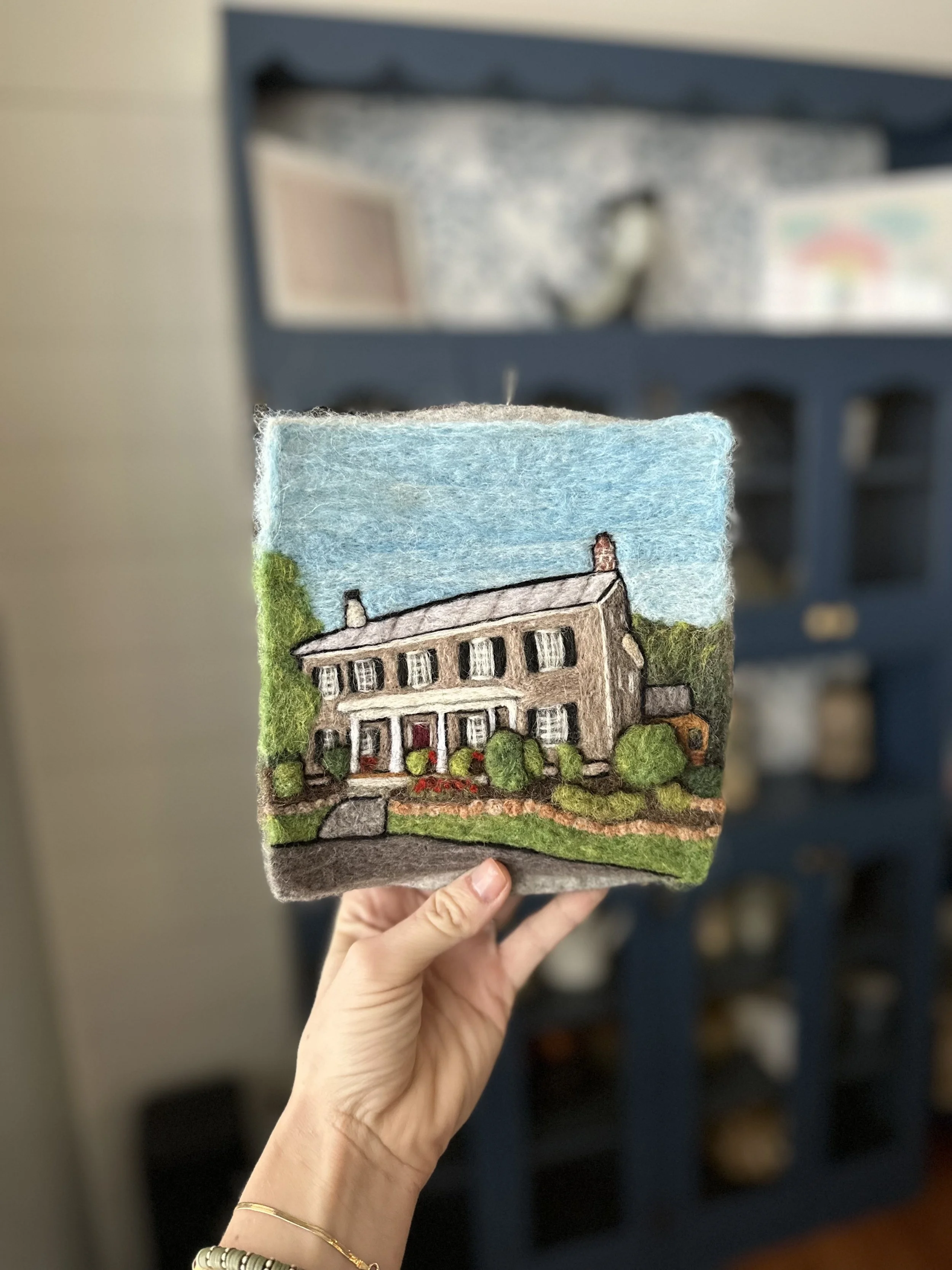 Close-up of a felt artwork featuring a multi-story house with a grass yard, bushes, and a blue sky background.