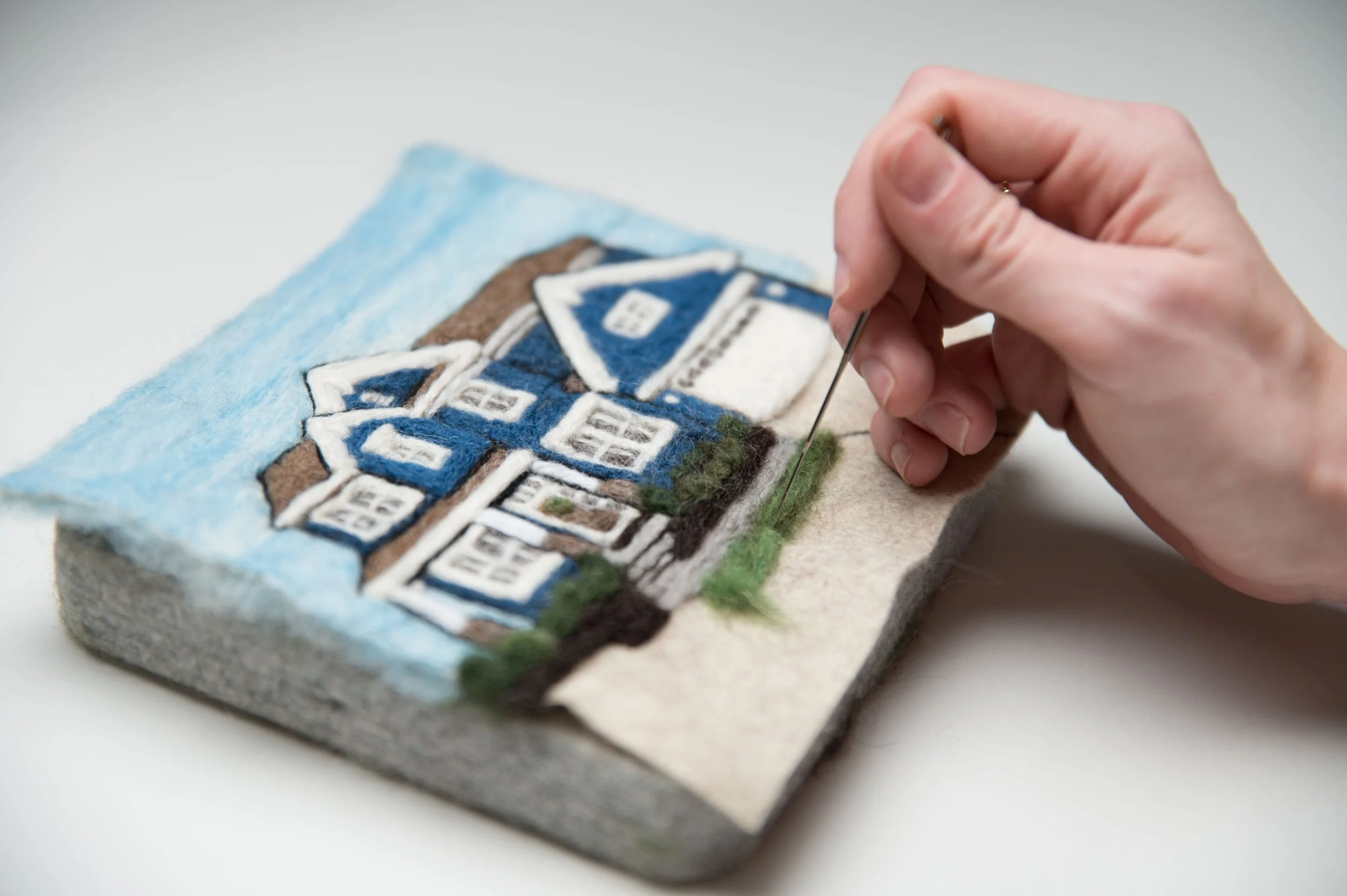 Close-up of a person sewing or embroidery on a felt fabric depicting a house with a blue roof, white windows, and brown walls.