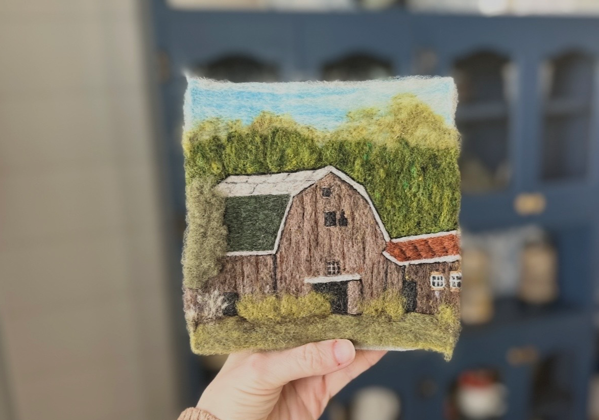 Felted artwork of a barn with trees and a blue sky in the background.