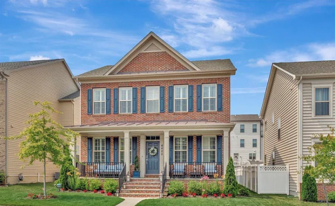 Two-story brick house with front porch, potted plants, and decorated door, surrounded by lawn and neighboring houses.