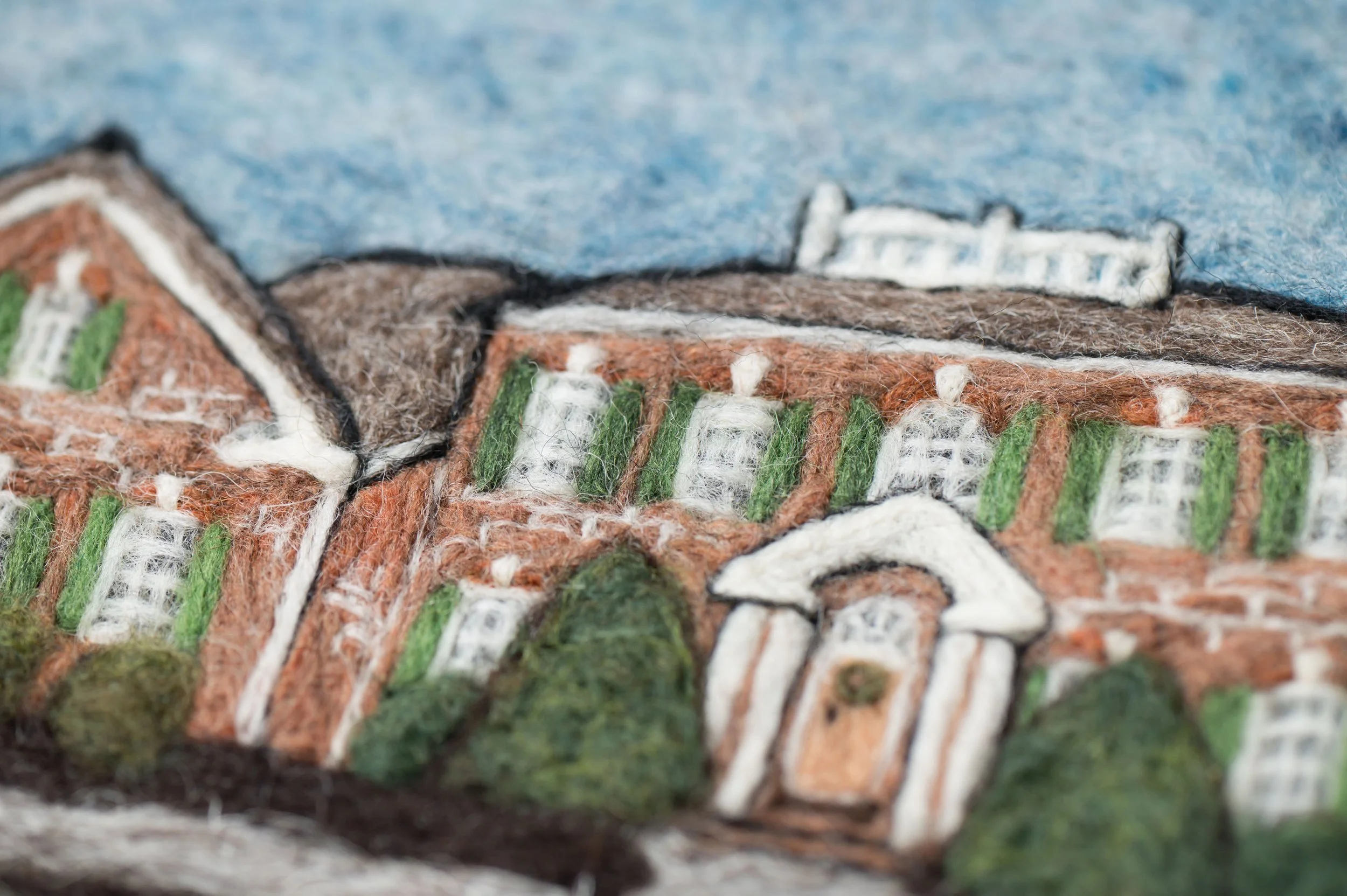 Close-up view of embroidered fabric depicting a landscape scene with trees, buildings, and a river, using various colored threads like green, brown, white, and blue.