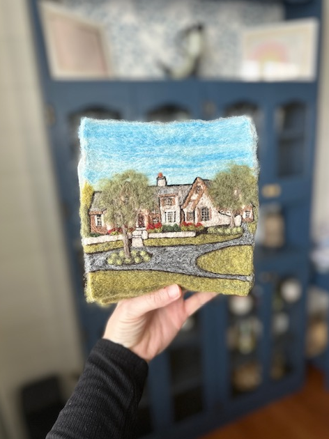 A piece of artwork depicting a house with trees, a driveway, and a cloudy sky, held in a person's hand indoors.