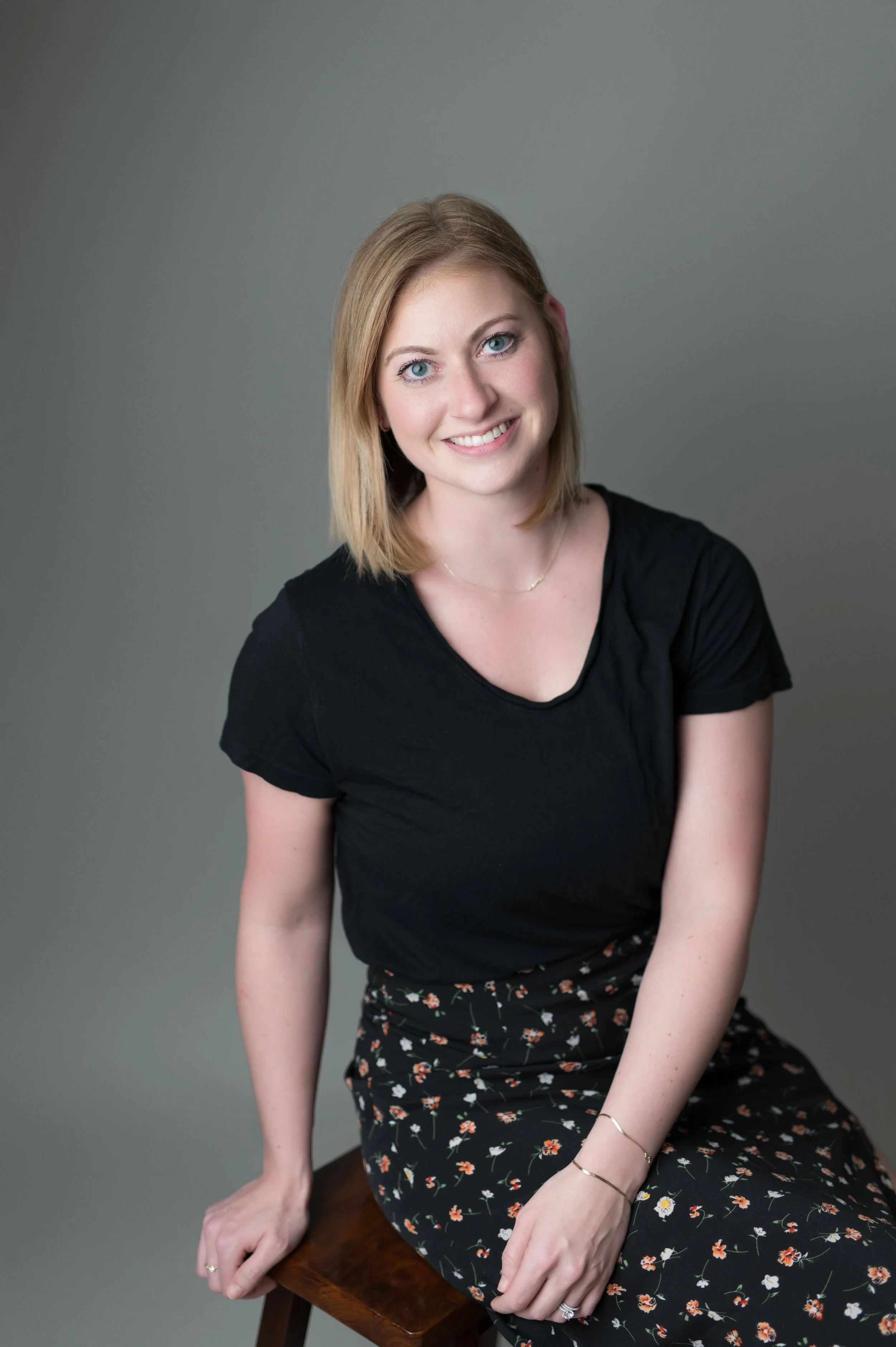 A young woman with blonde hair and blue eyes smiling, wearing a black t-shirt and a floral skirt, sitting on a wooden stool against a gray background.