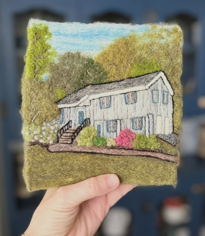 A piece of felt artwork depicting a white house with a staircase, surrounded by green trees and bushes, under a blue sky.
