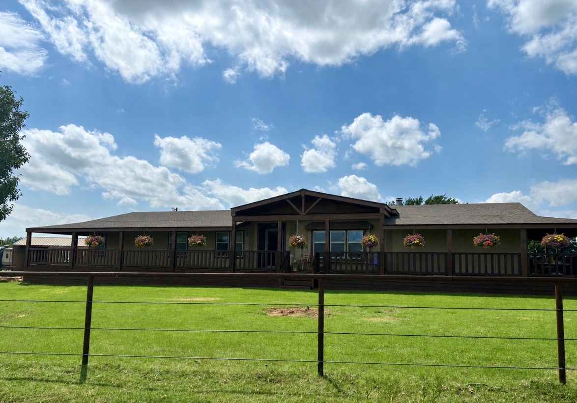 A single-story house with a large front porch decorated with hanging flower baskets. The house is surrounded by a grassy lawn and a black wire fence, with a bright blue sky and scattered clouds overhead.