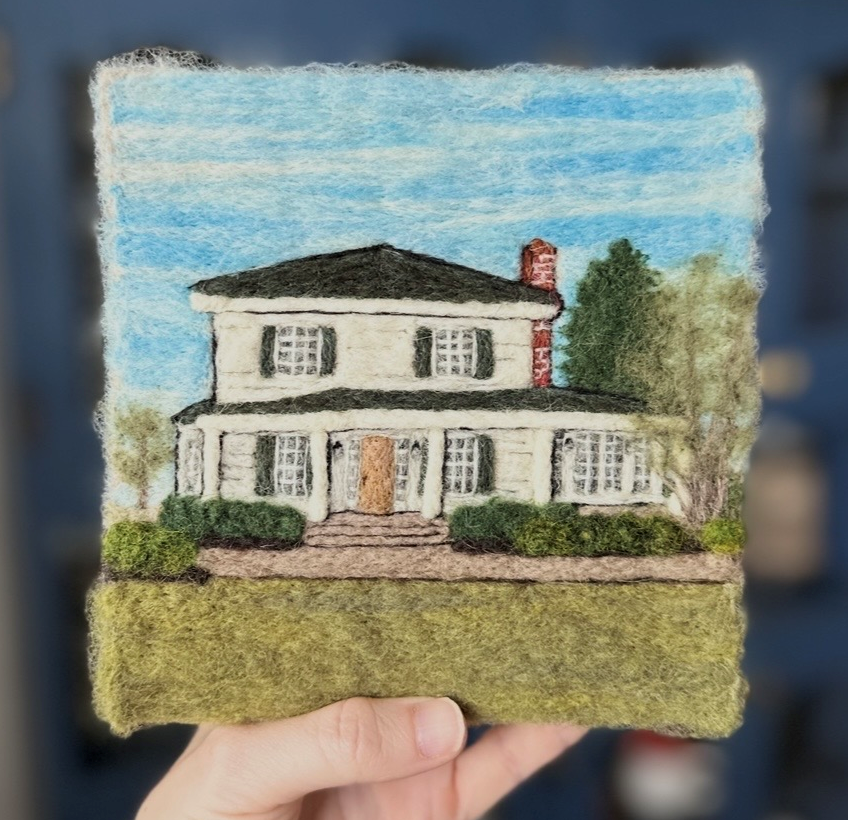 Felt art depicting a two-story house with a black roof, white walls, multiple windows with black shutters, a front porch with steps, green bushes, and trees in the background under a blue sky.
