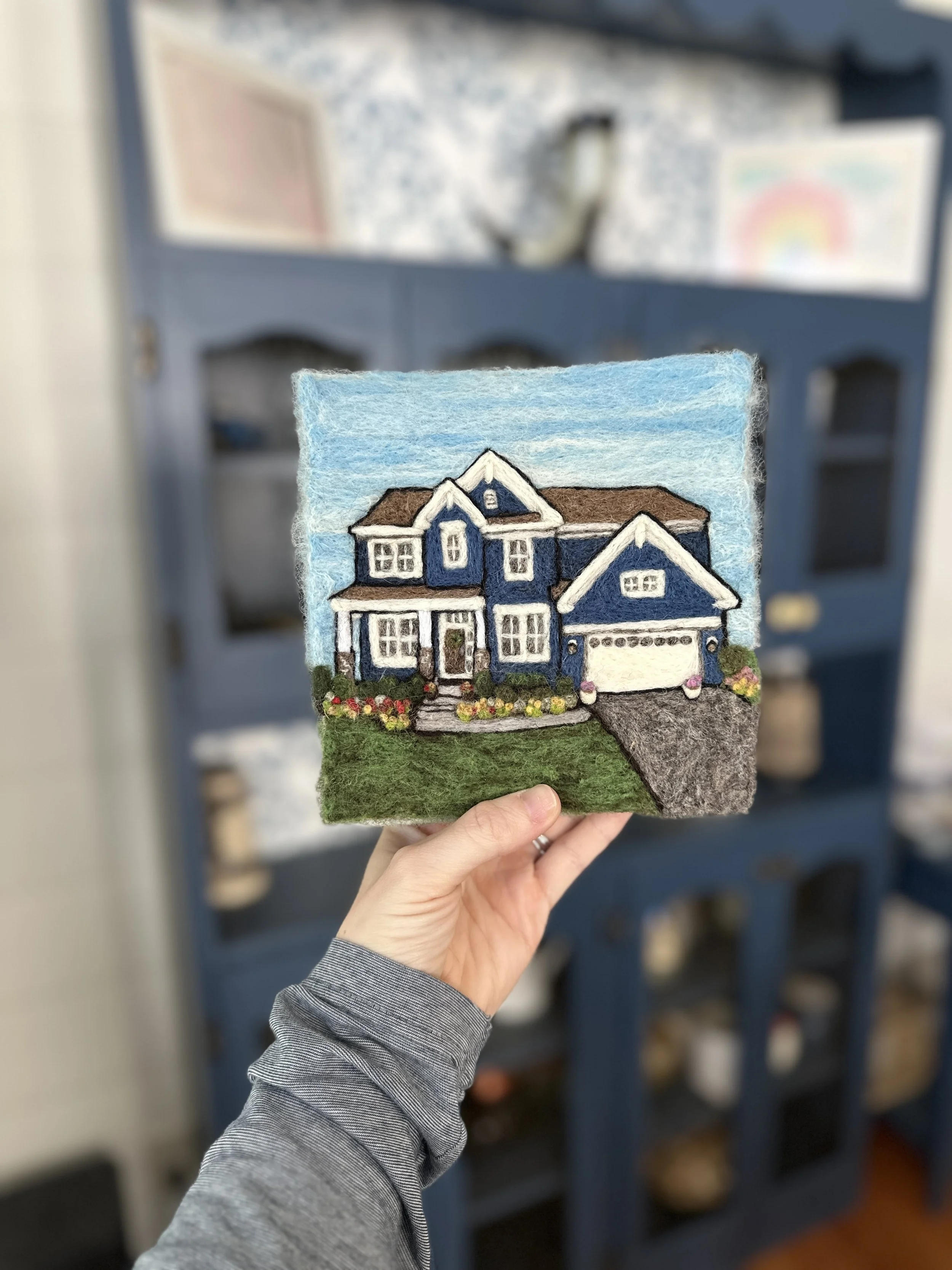 Felt artwork of a blue house with white trim and a front yard with flowers, held in hand in front of a blue bookshelf with various items.