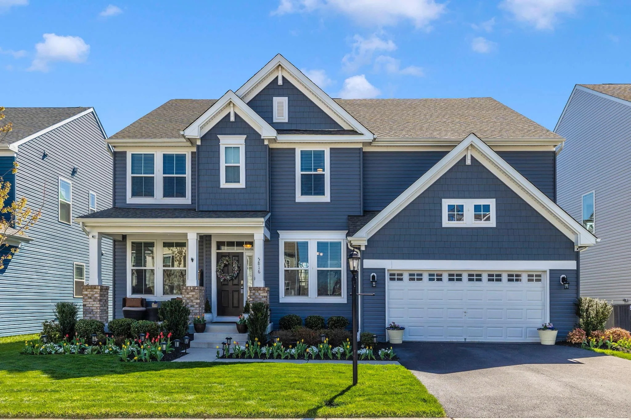 Front view of a two-story blue house with a white garage door, front porch, and well-maintained lawn and garden with colorful flowers and bushes.