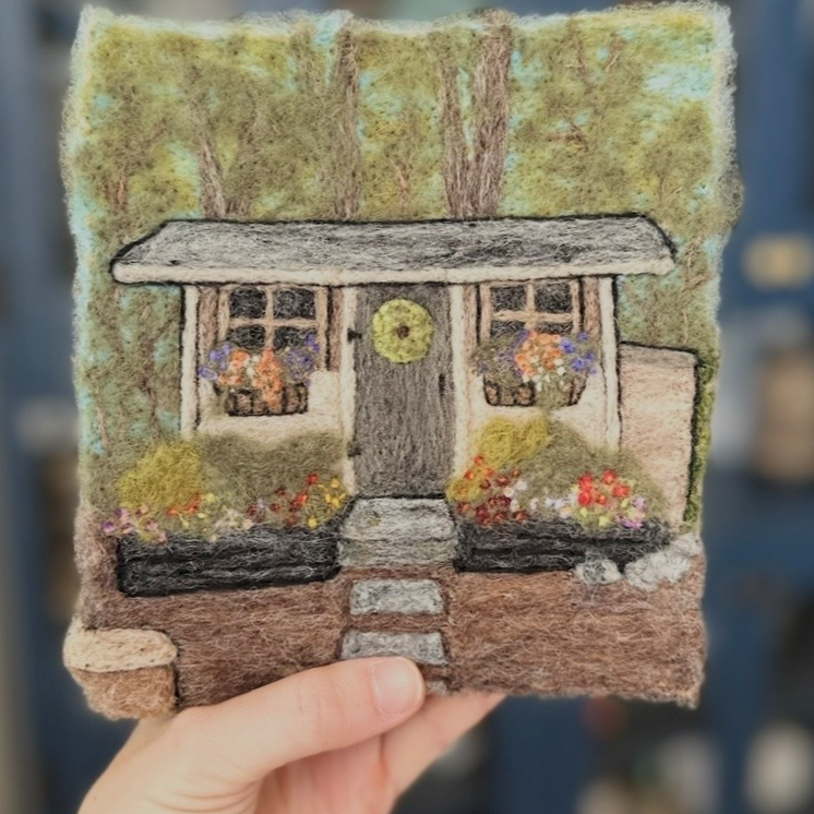 A hand holding a piece of artwork depicting a cozy house with a front door, windows with flower boxes, and a garden, set against a background of trees.