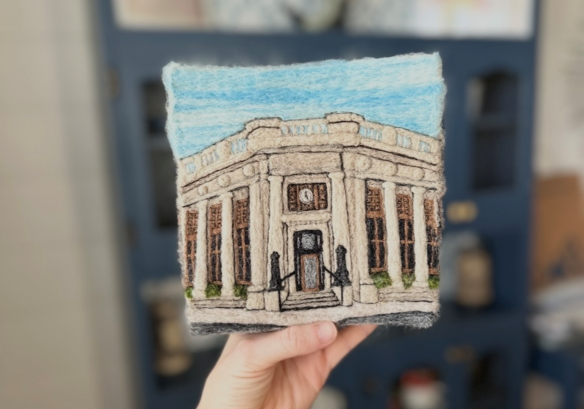 Felt hand-stitched artwork of a historic building with columns, a clock, and steps, held in a person's hand.