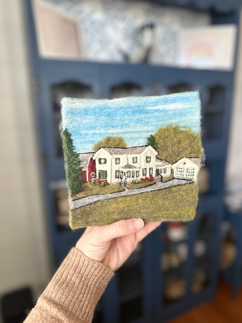 Hand holding a felted artwork of a house with trees, a driveway, and a blue sky in front of a blue cabinet shelf.