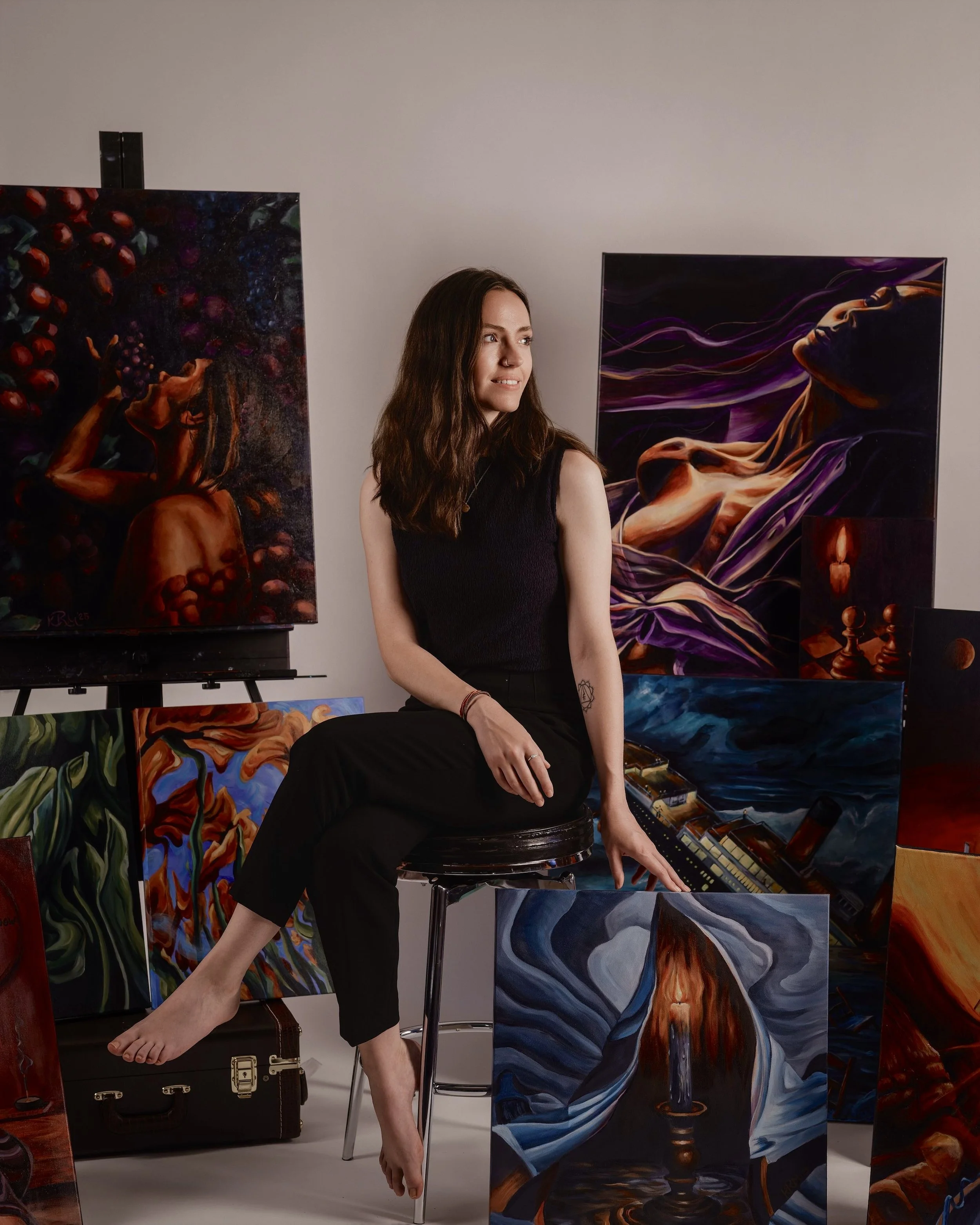 Young woman with long brown hair sitting on a stool in an art studio, surrounded by colorful paintings of surrealistic and abstract scenes.