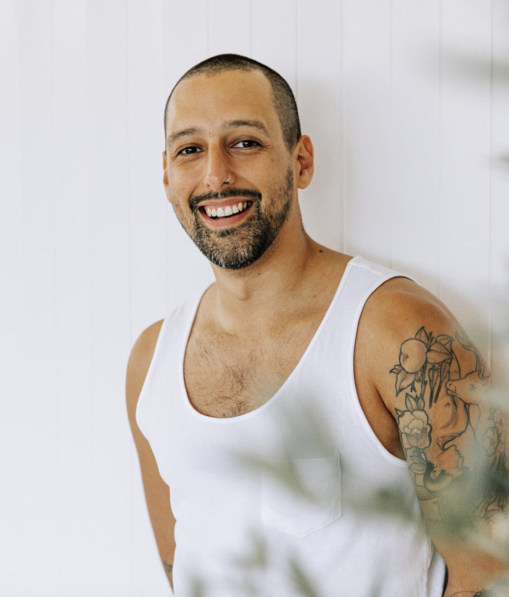 A smiling man with a shaved head, beard, and tattooed arm, wearing a white tank top, standing against a white wall.