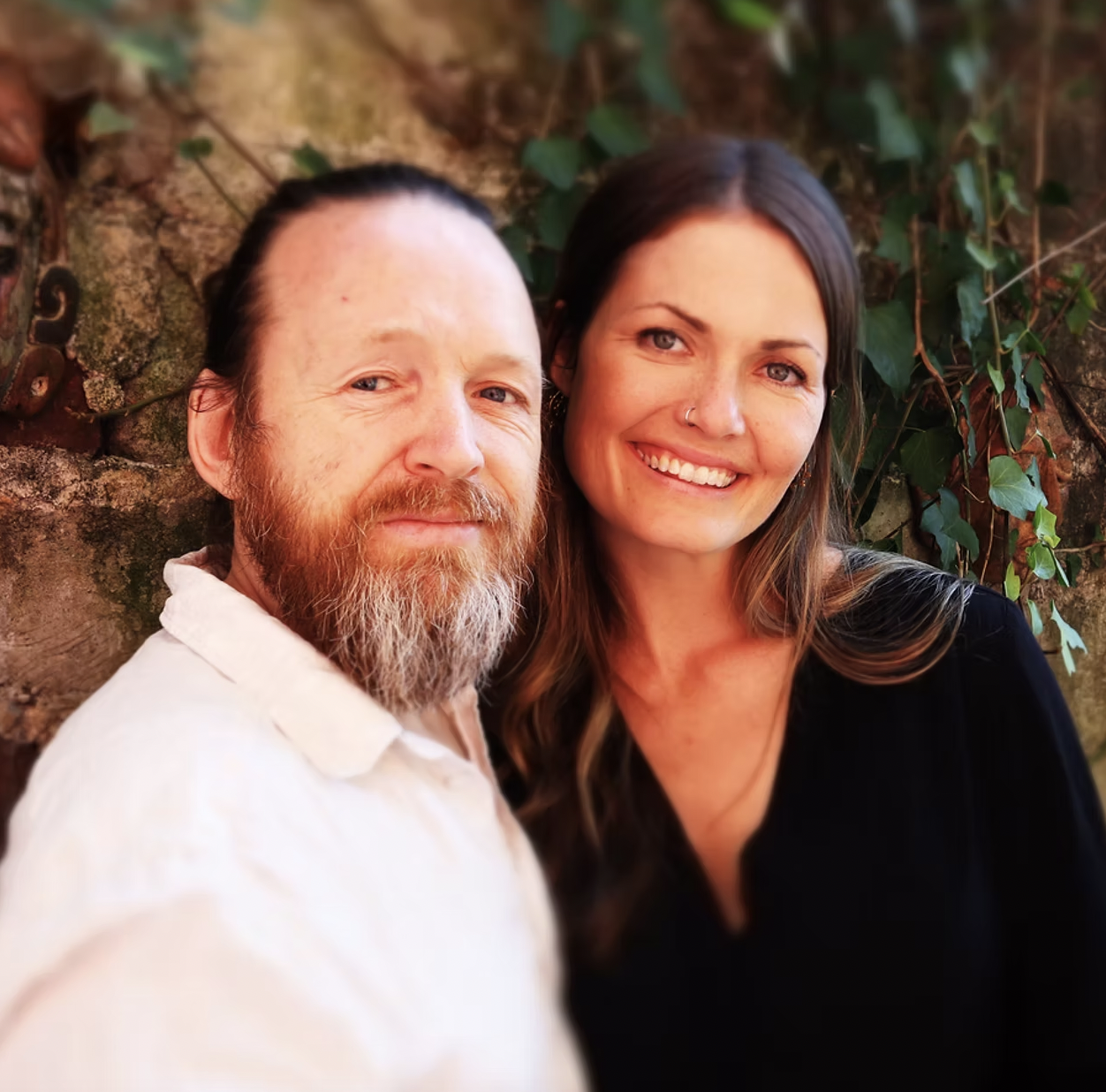 A man with a beard and long hair and a woman with long brown hair smiling for a selfie in front of a brick wall covered with ivy.