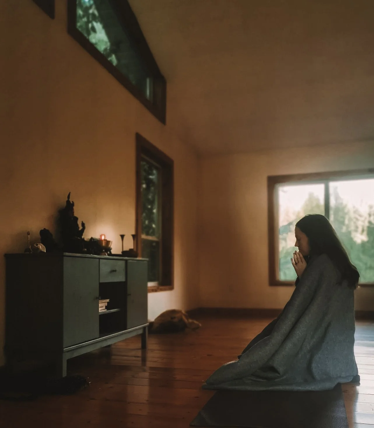 Woman kneeling on a yoga mat in a softly lit room, facing a small altar with a statue and candles, with windows showing green trees outside.