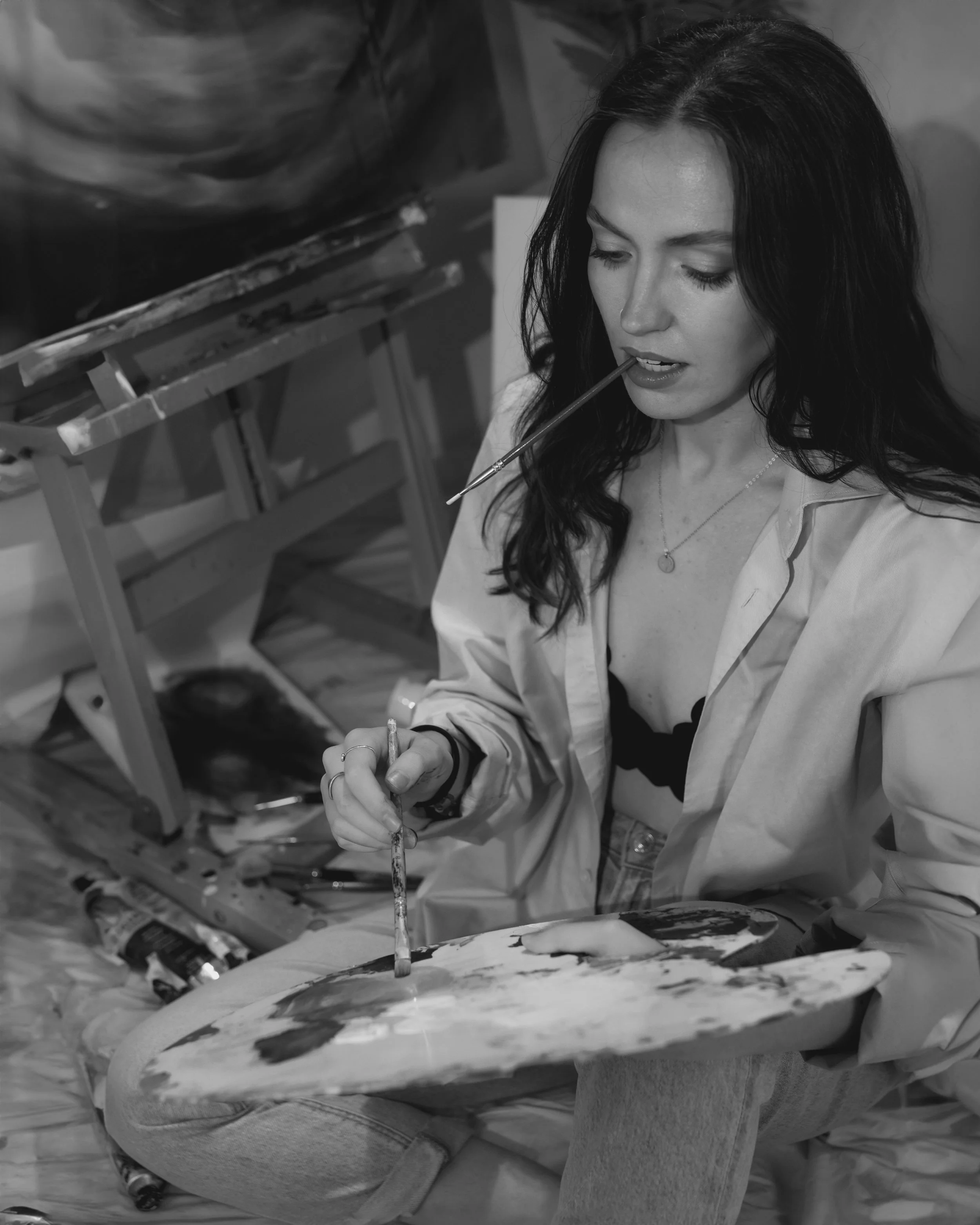 A woman sitting cross-legged, holding a painter's palette and using a brush to paint on a canvas. She is biting the end of a paintbrush.
