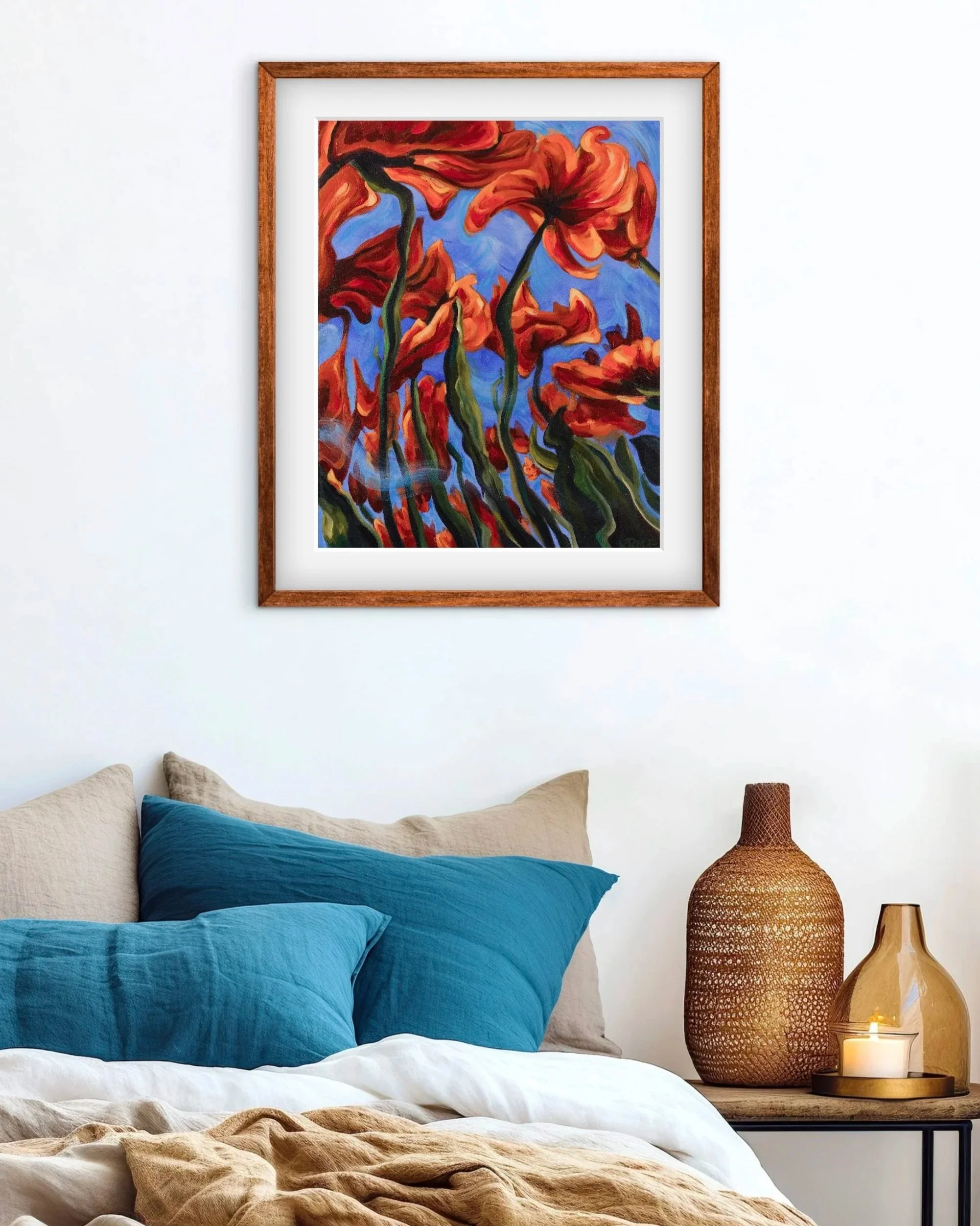 A framed painting of abstract orange flowers on a blue background hanging above a bed with blue pillows and beige bedding. To the right, a bedside table with two decorative vases and a lit candle.