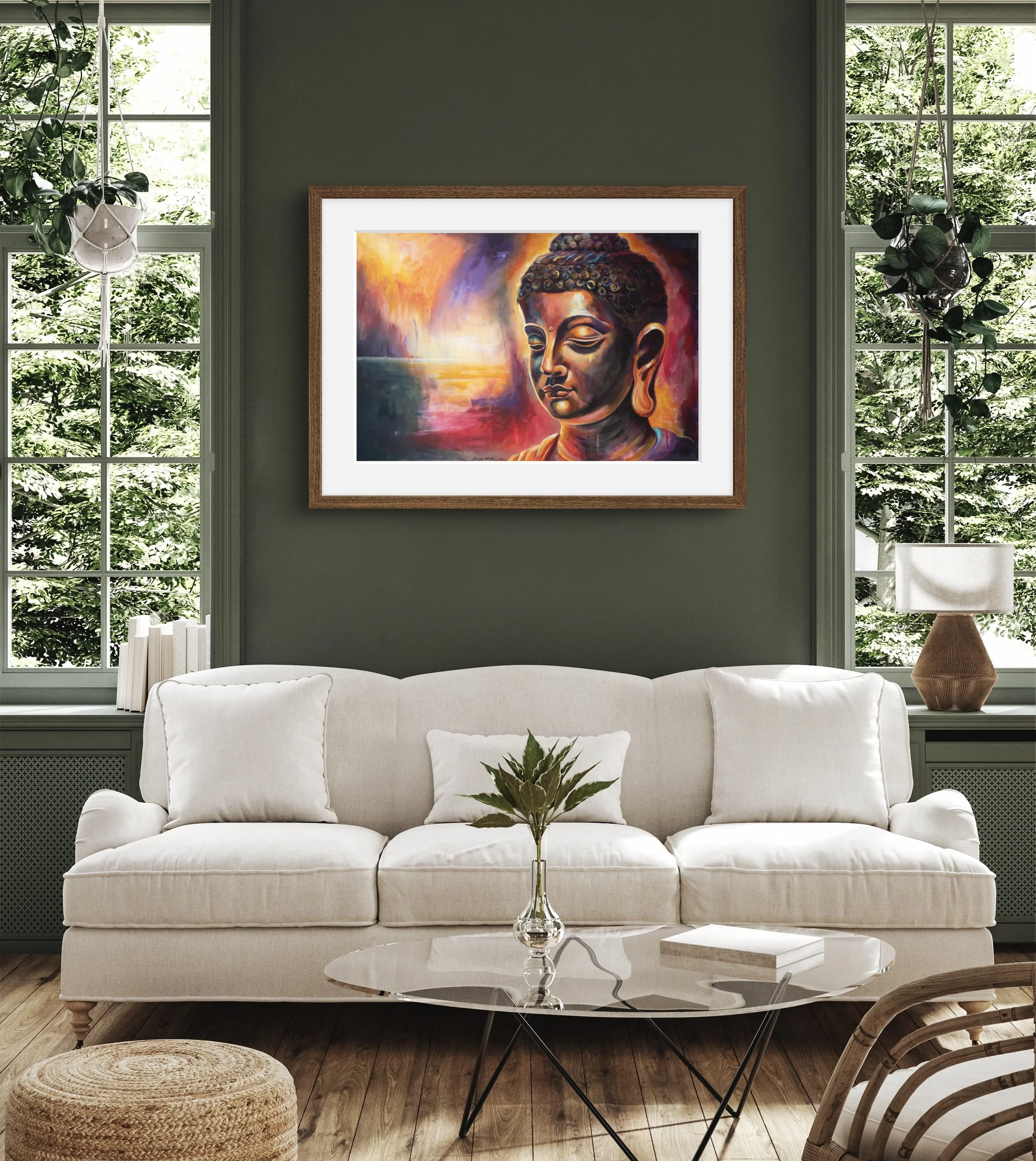 A living room featuring a white sofa with cushions, a glass coffee table with a book and a vase of leaves, large windows with greenery outside, and a framed colorful painting of Buddha hanging on a dark green wall.