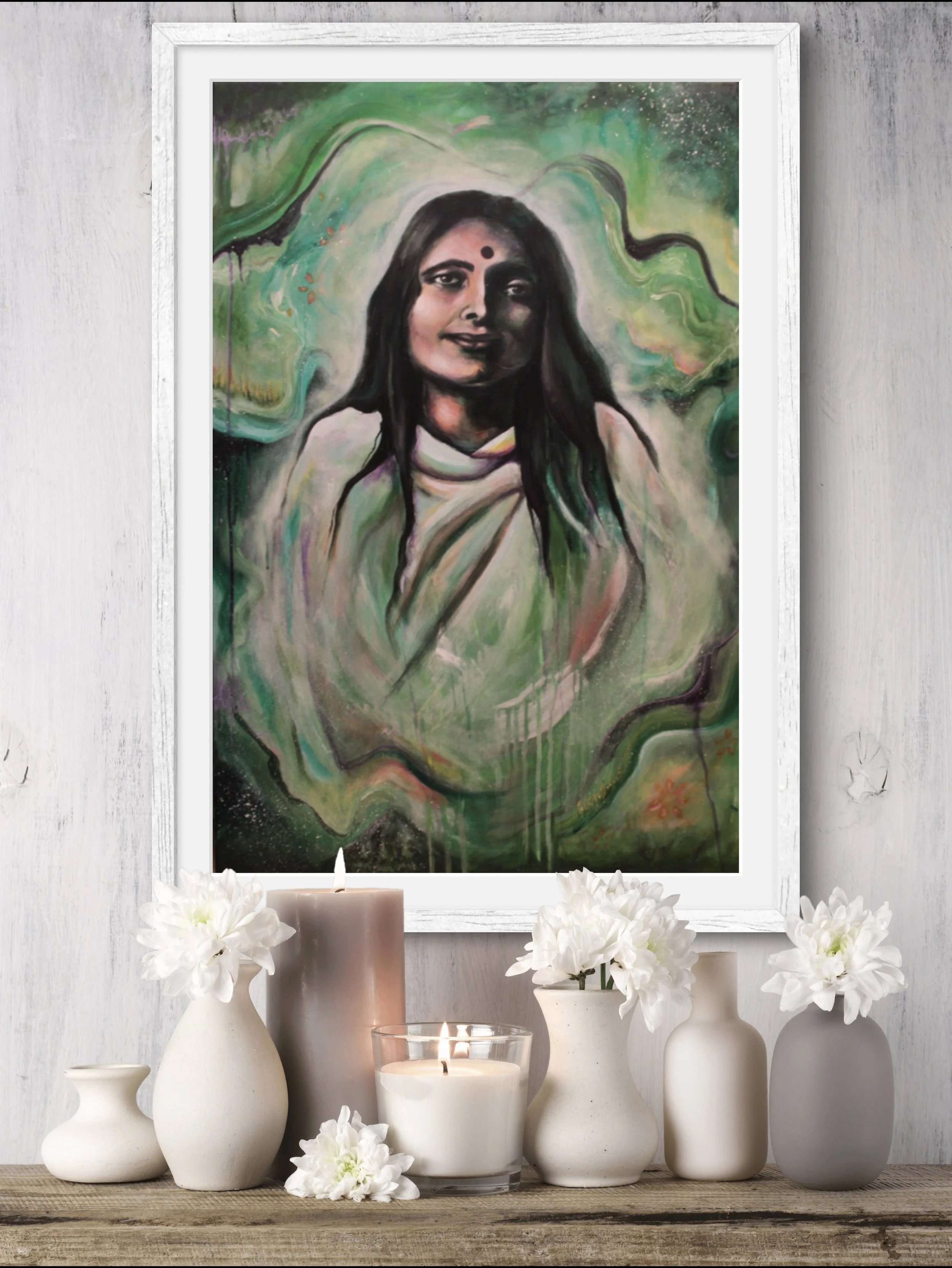 A framed portrait of a woman with dark hair, a bindi on her forehead, and a gentle smile, set against a colorful abstract background with greens and blacks. Below the portrait, there are white vases with flowers, candles, and a wooden surface.