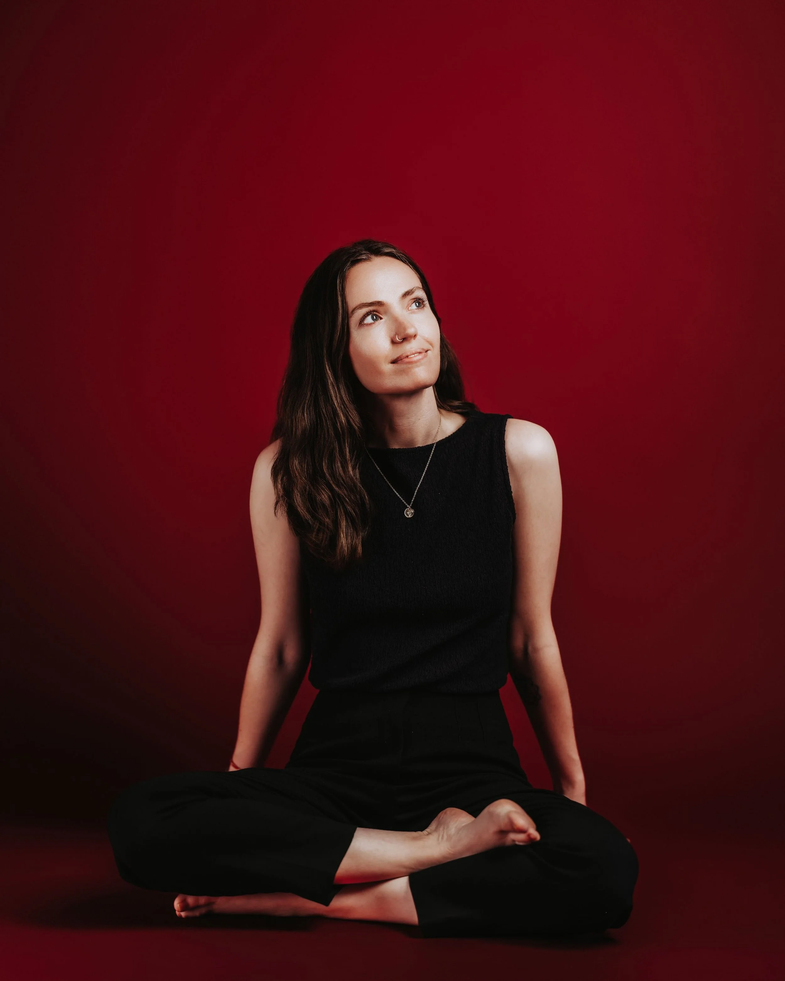 A woman sitting cross-legged on a dark red background, looking upward. She has long wavy brown hair, wearing a black sleeveless top and black pants, with a silver necklace.