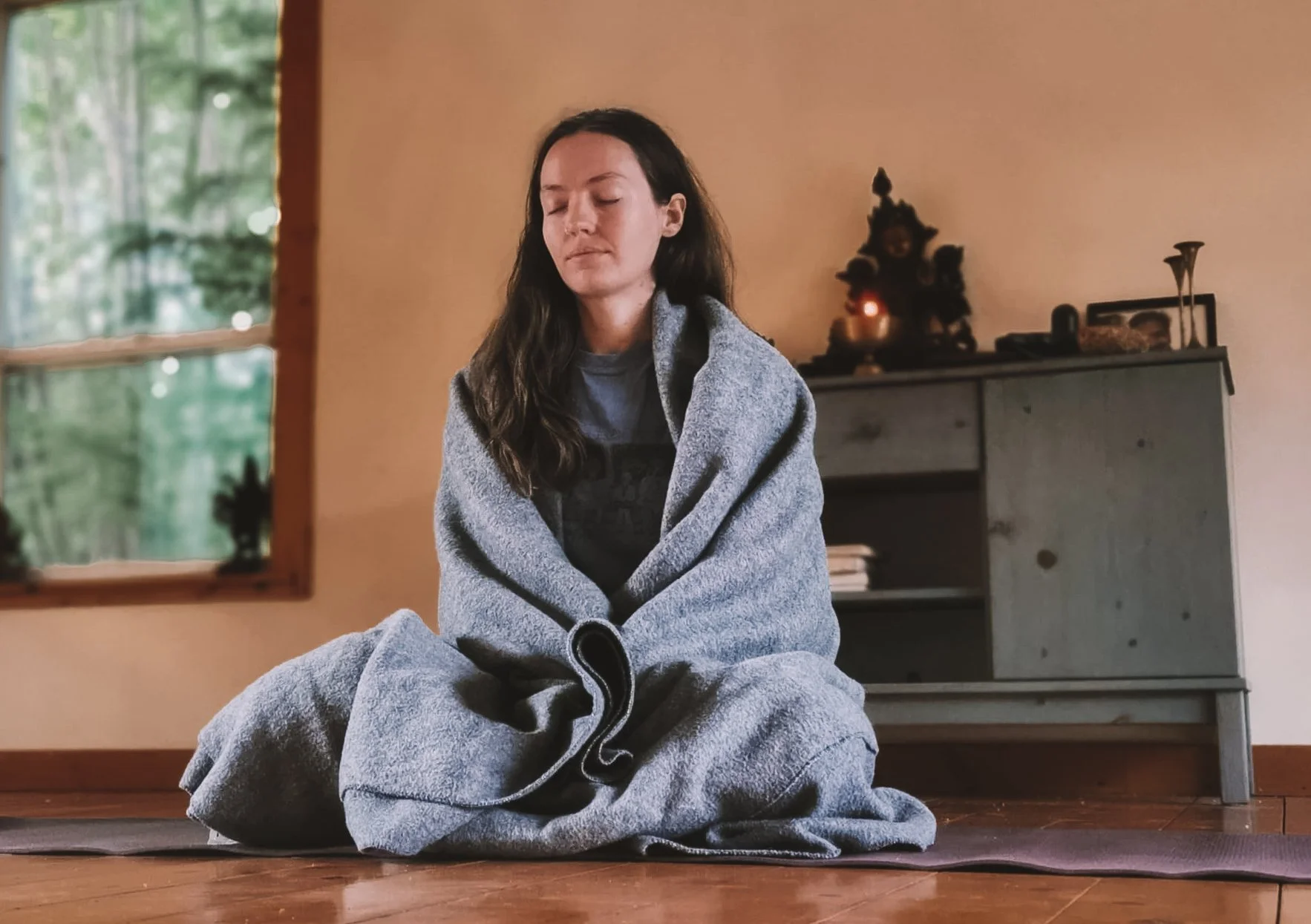 A woman sitting on a yoga mat indoors, wrapped in a gray blanket with her eyes closed, in a meditative pose.
