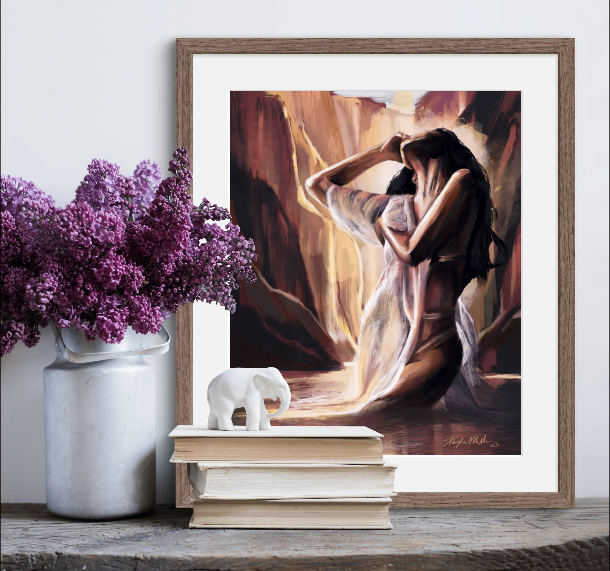 A still life with a framed painting of a woman with long dark hair, standing between rocky cliffs with her arms raised and head bowed, next to a white vase of purple flowers, a small white elephant figurine, and a stack of beige and white books on a rustic wooden surface.