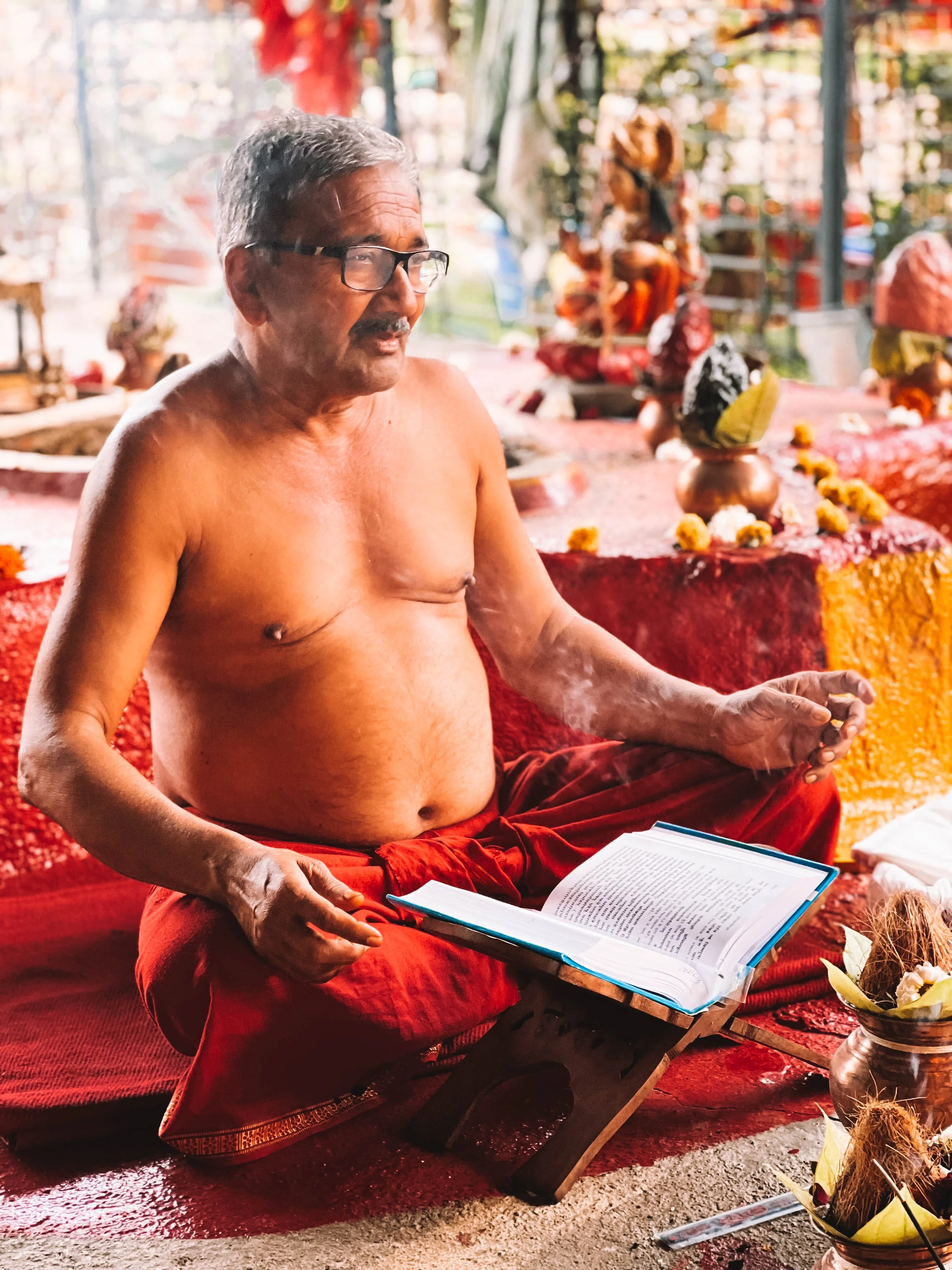An elderly man sits cross-legged on the ground, shirtless, reading a book placed on a wooden stand. He has gray hair, wears glasses, and is dressed in red traditional attire. In the background, there are temple statues, flower garlands, and offerings, indicating a religious or spiritual setting.