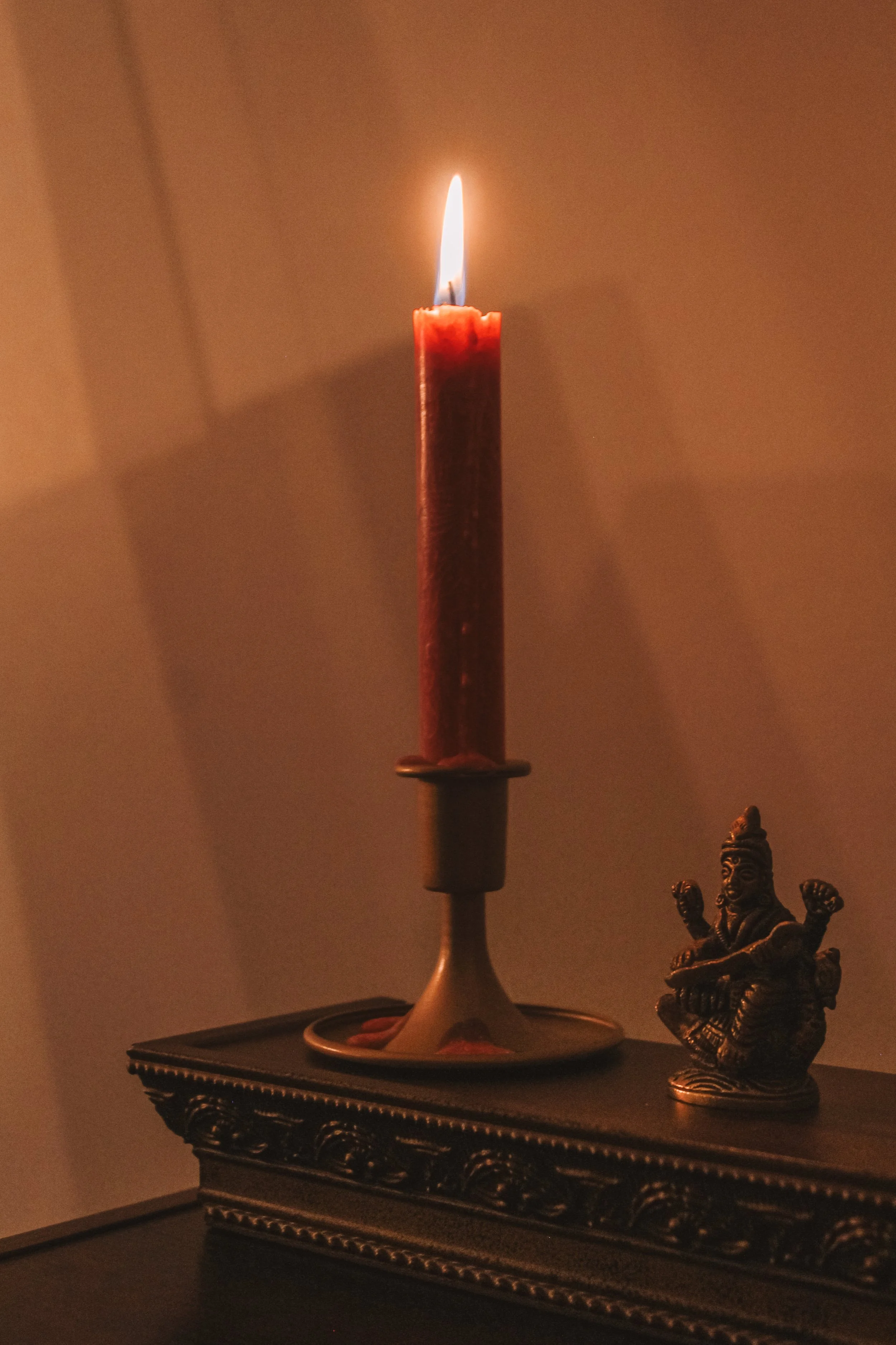 A lit red candle on a brass candle holder, placed on a decorative black surface with a small brass figurine of a seated deity nearby.