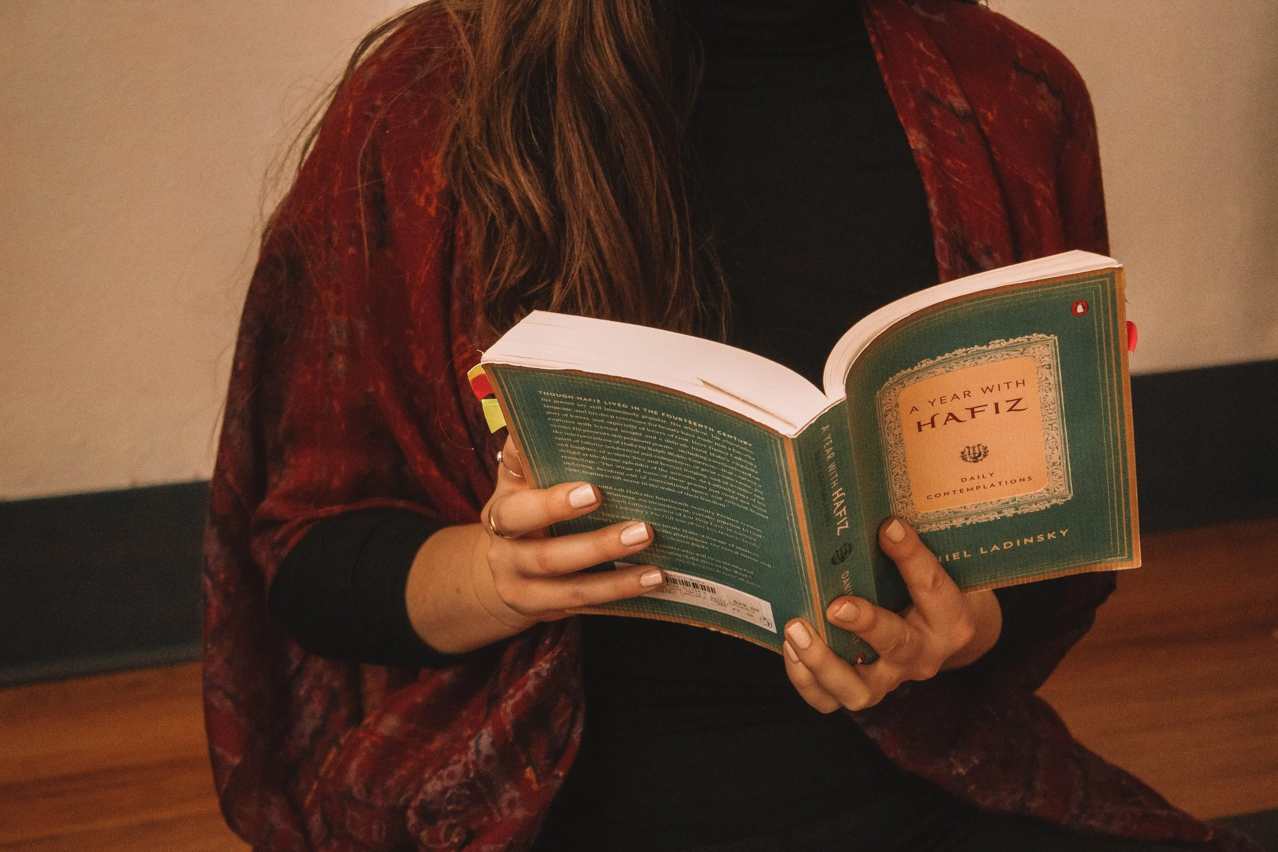 A person reading a book titled "A Year with Hafiz" by Daniel Ladinsky, seated indoors with a wooden floor, wearing a black top and a red patterned shawl.