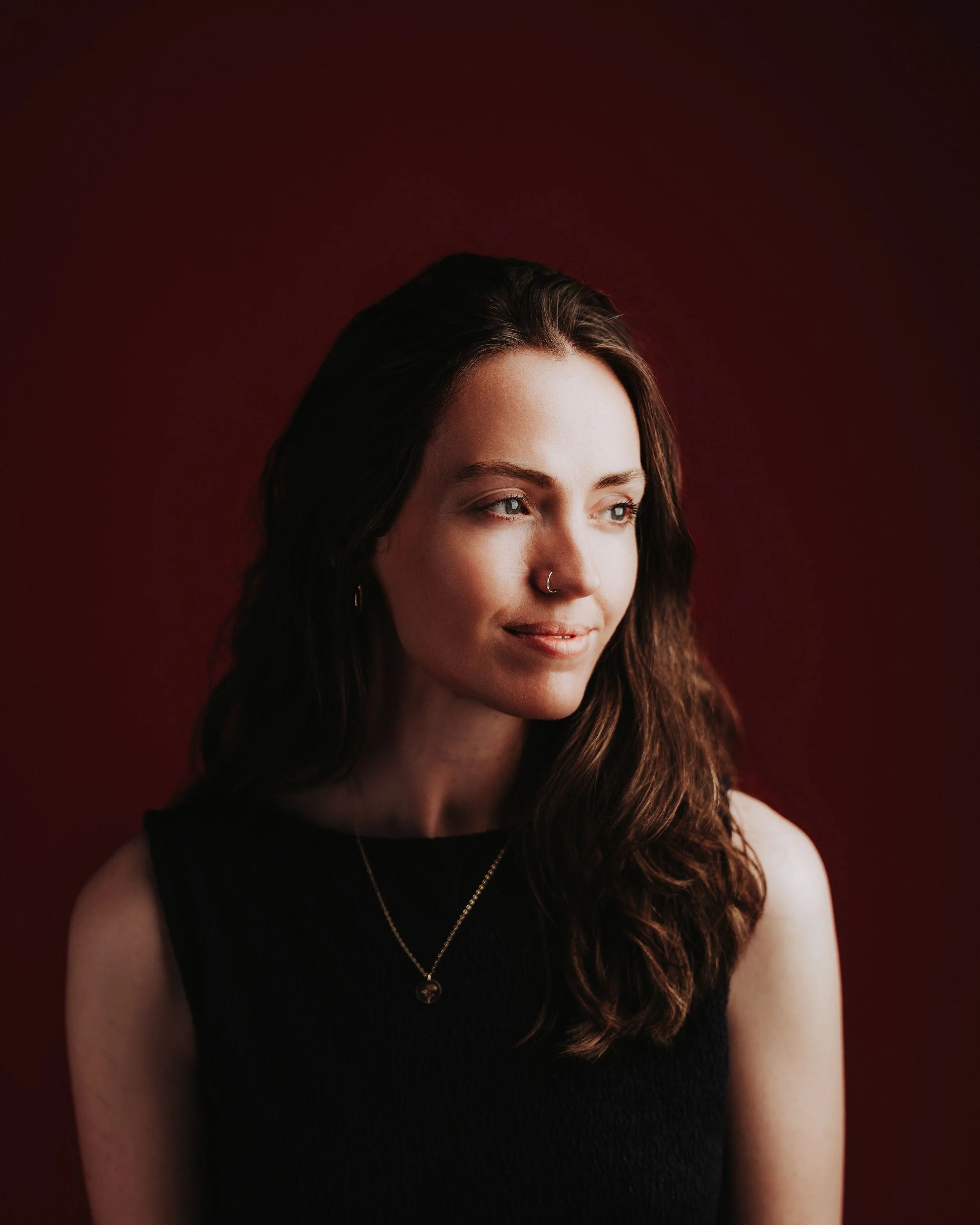 A woman with long dark hair, blue eyes, wearing a black sleeveless top, a silver necklace, and a nose ring, looking slightly to the side against a dark red background.