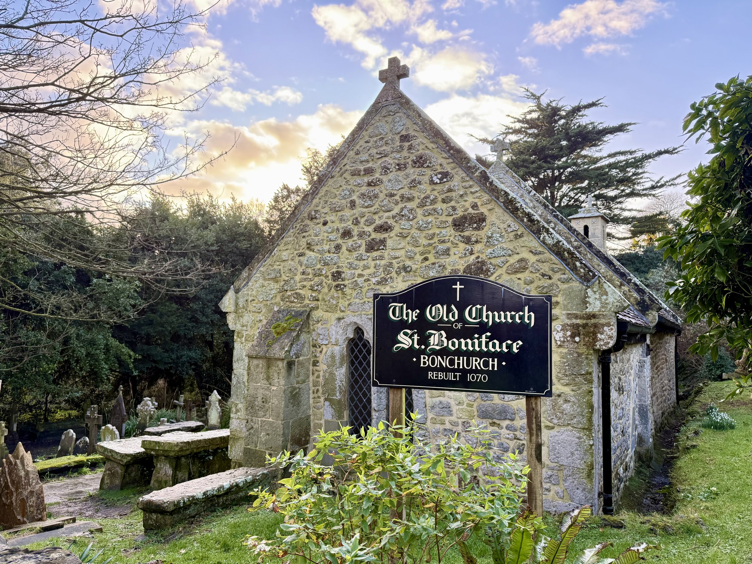Old stone church with sign reading 'The Old Church of St. Boniface, Bonchurch, Rebuilt 1070,' surrounded by trees and gravestones, under a partly cloudy sky.