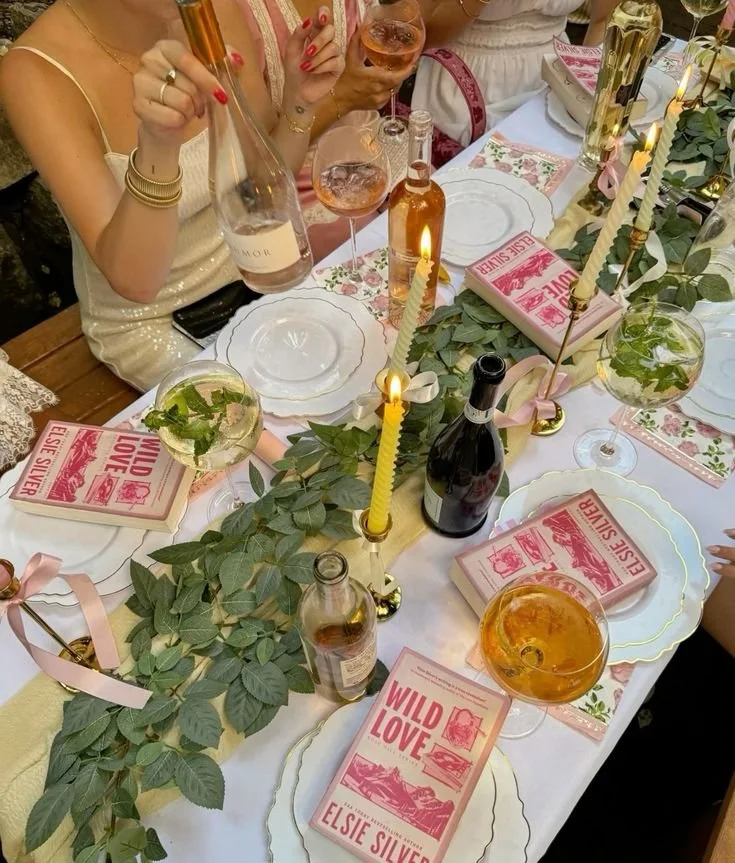 A table decorated for a gathering with a green leafy table runner, pink candles, white plates, pink books titled 'Wild Love,' and various glasses of rosé wine.