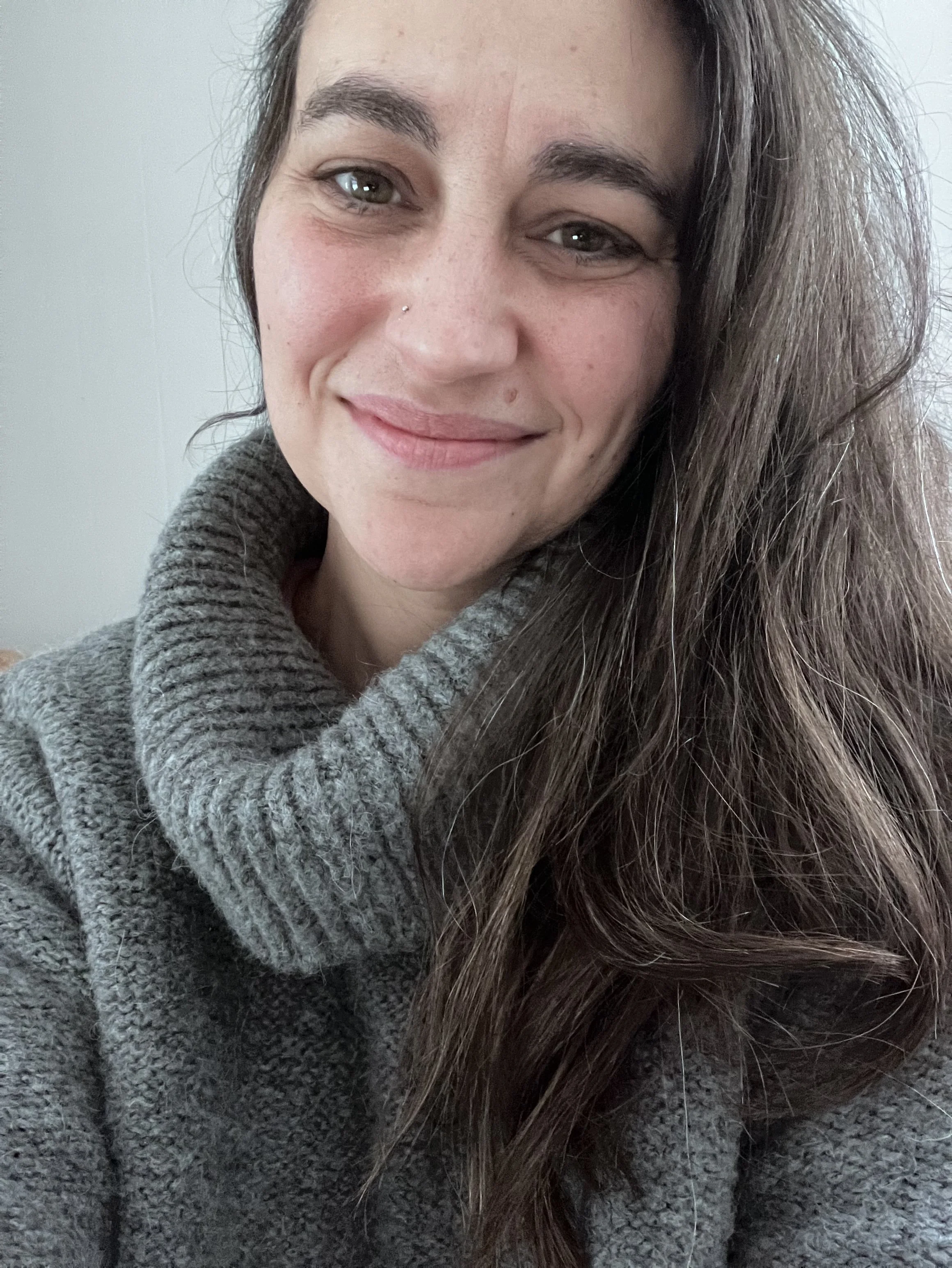 Close-up of a smiling woman with long brown hair, wearing a gray turtleneck sweater, against a plain background.