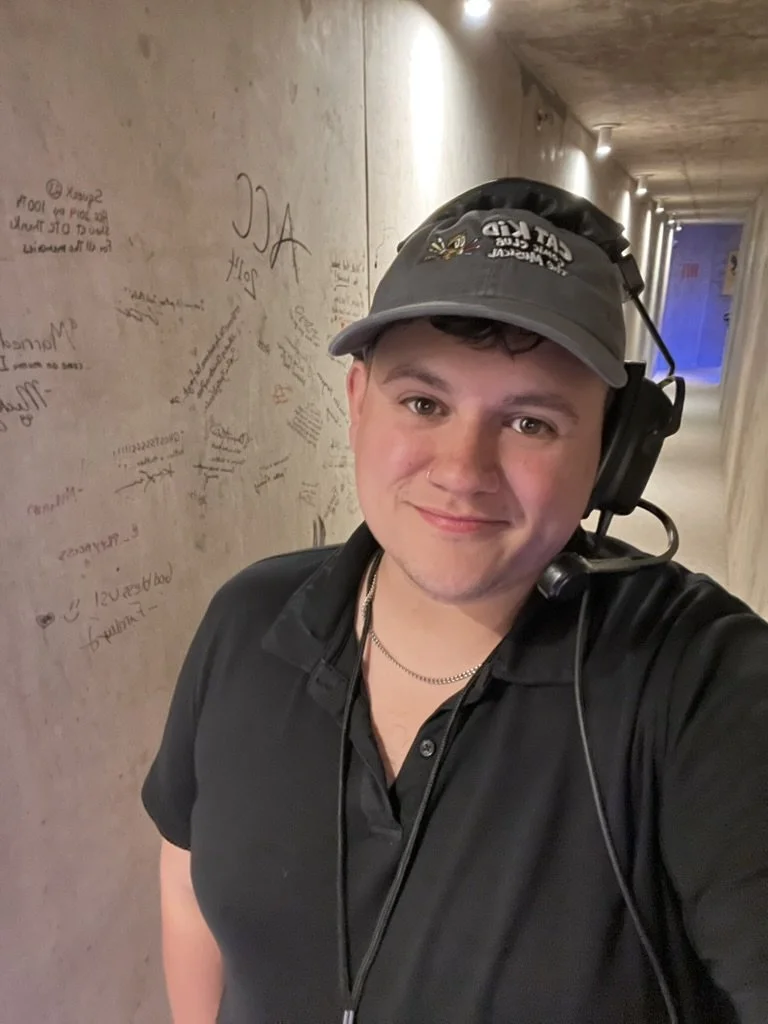 Colton Robertson smiling in a backstage hallway wearing a comms headset and a Cat Kid Comic Club: The Musical cap