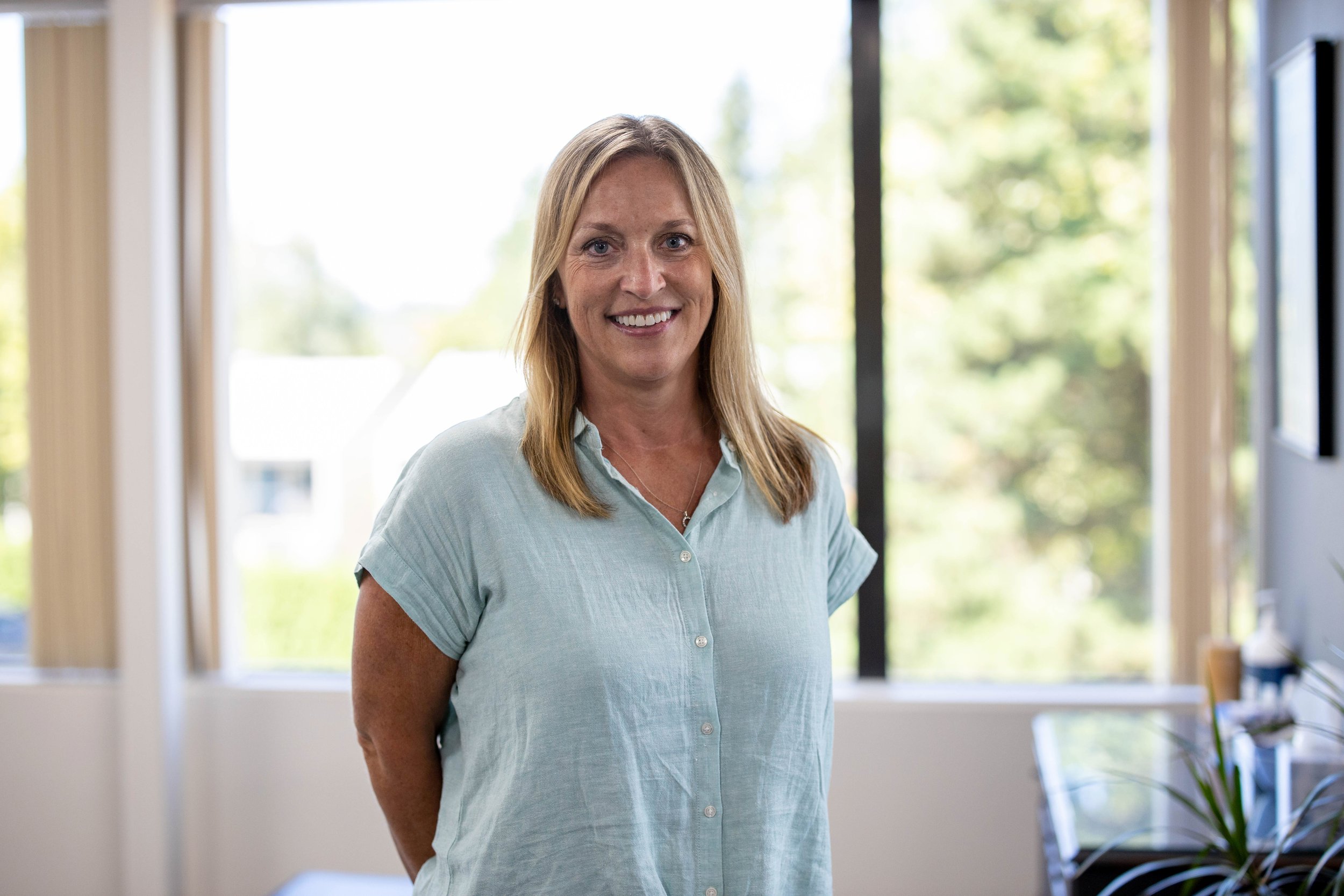 Smiling woman with shoulder-length hair standing in a bright office with large windows and green trees outside.