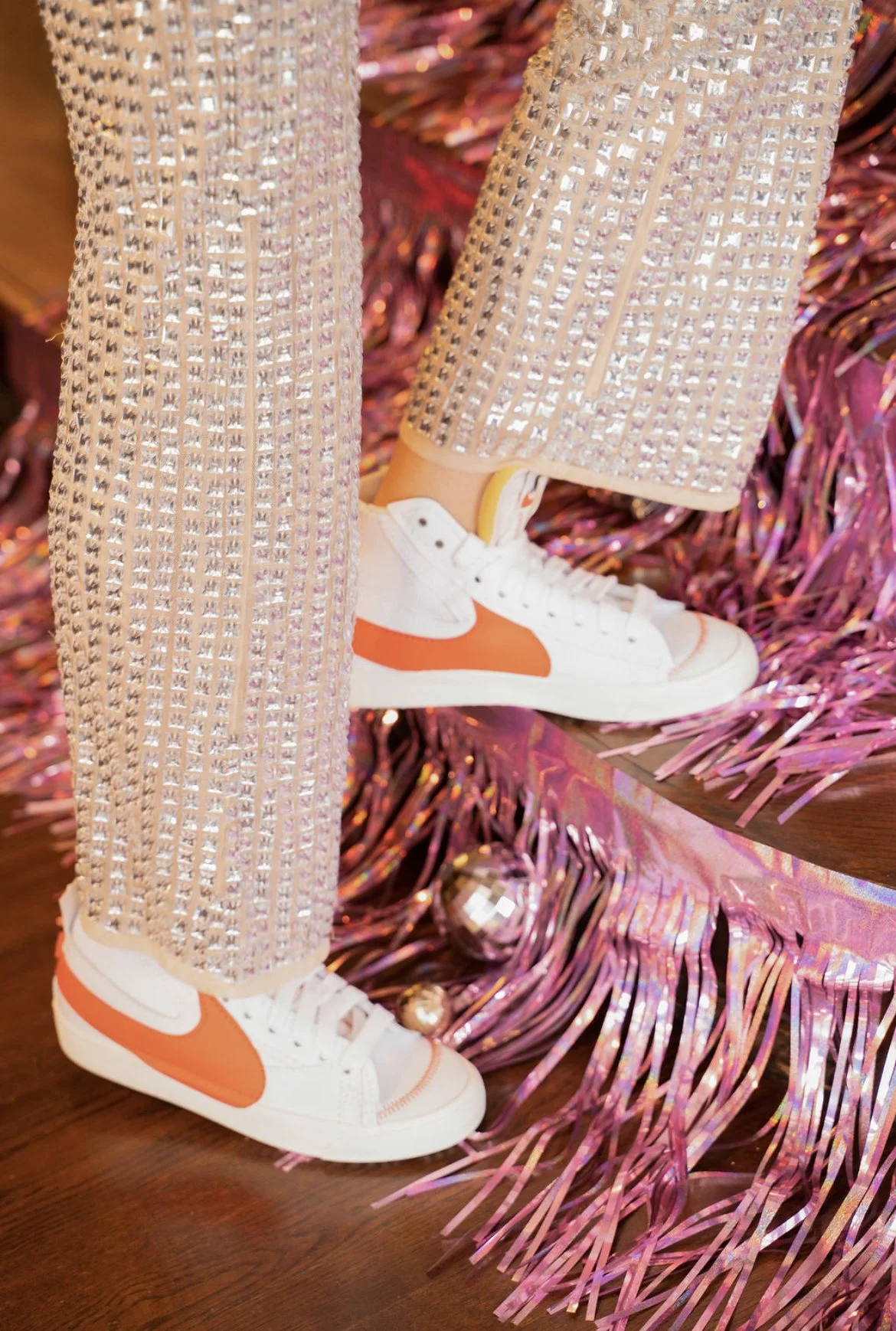 Person wearing white sneakers with orange accents and pink, sequined pants standing on pink tinsel decorations.